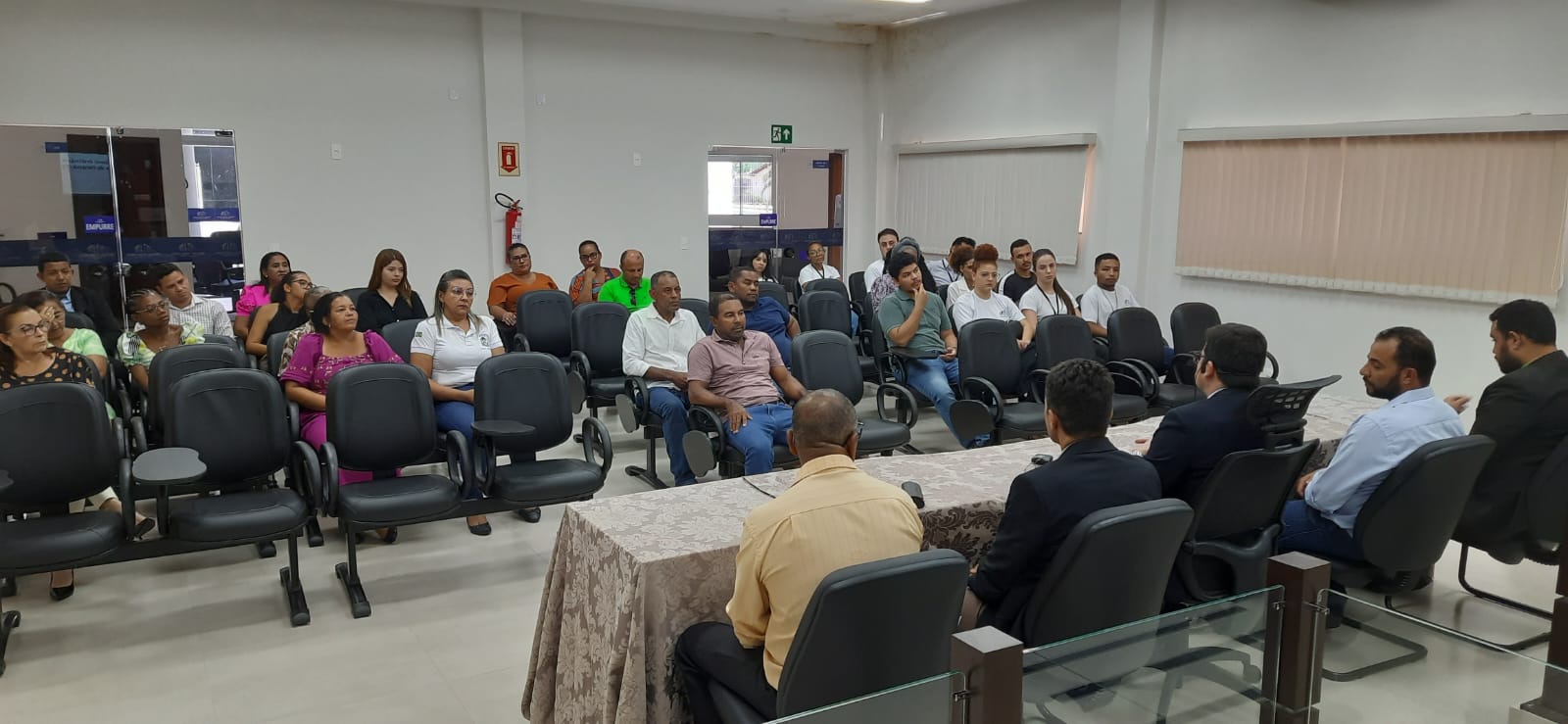 Fotografia colorida que mostra cinco homens sentados à mesa conversando. Eles são observados por uma plateia