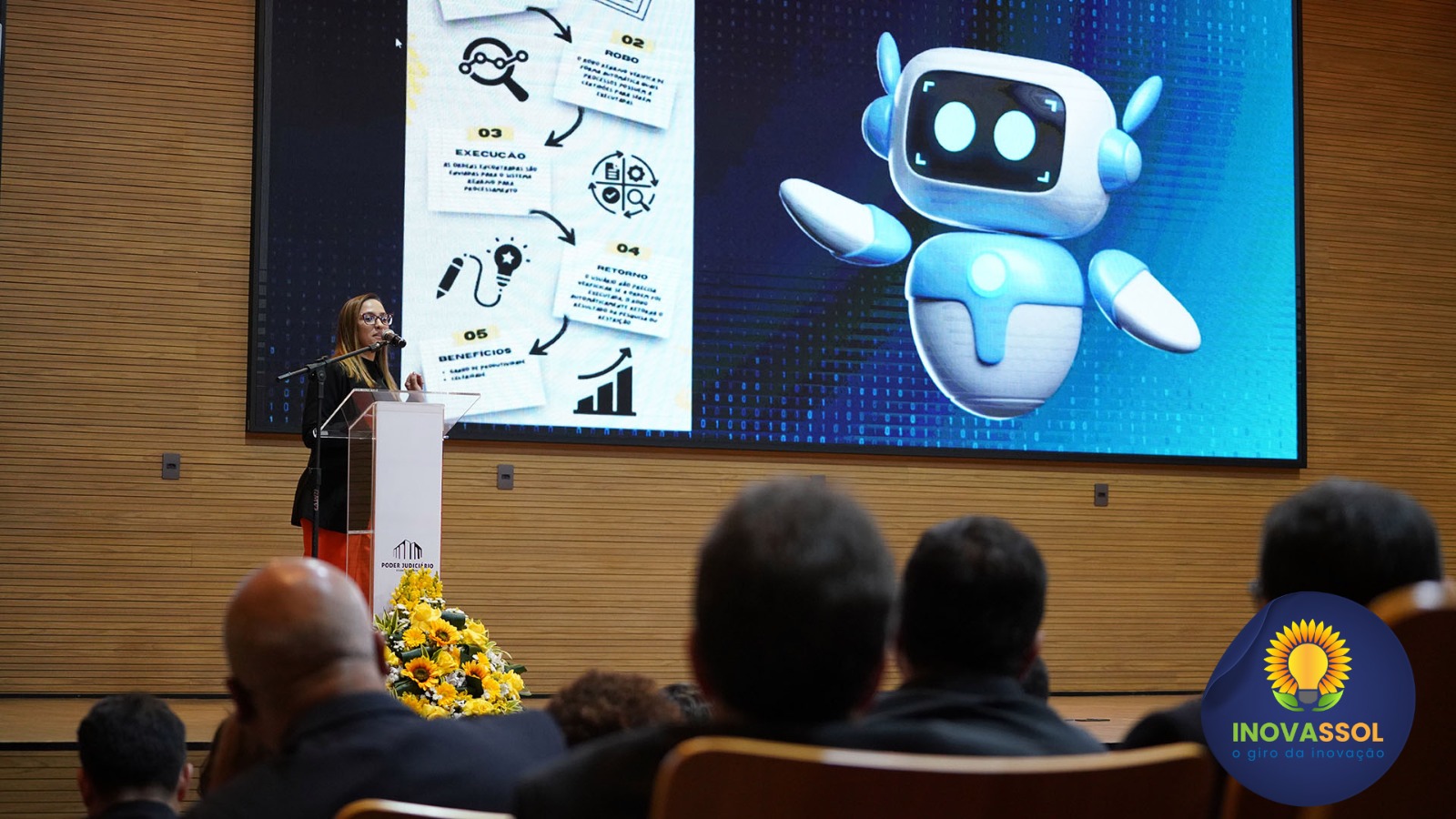 Mulher vestida com roupa vermelha e preto está atrás do púlpito e ao fundo um telão com imagem de robô; à frente plateia assiste sentada em cadeiras