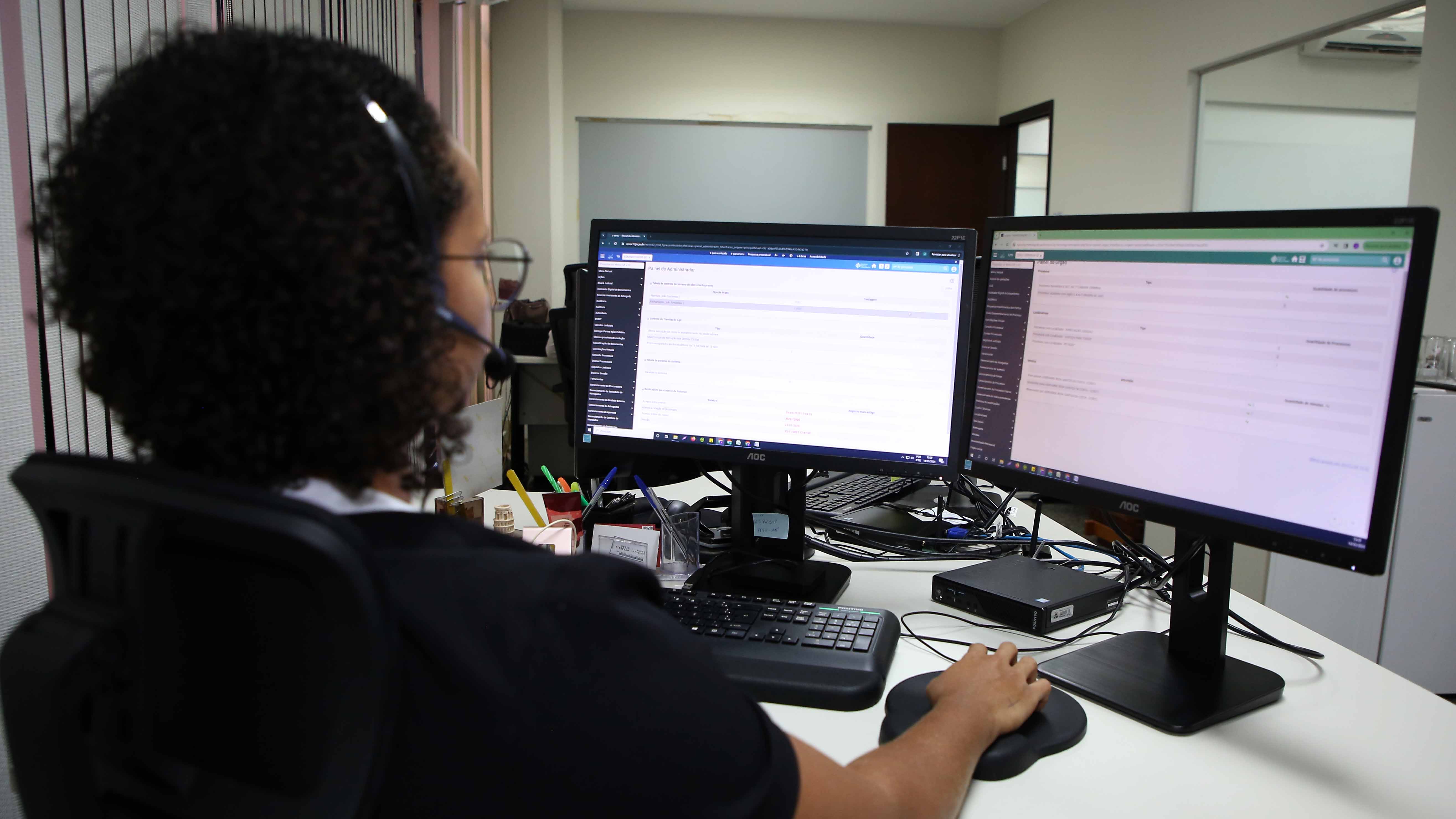 Moça de camiseta preta e cabelos curtos cacheados, sentada de costas enquanto opera um computador de duas telas, numa sala de paredes brancas