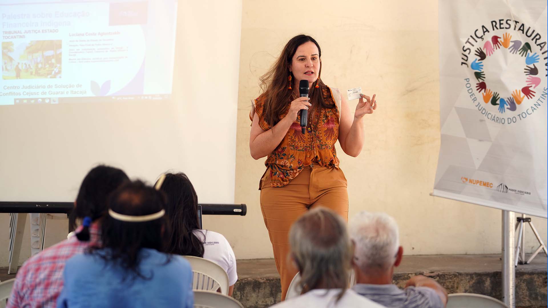 Foto de mulher branca, de cabelos castanhos, longos, soltos, vestida de calça e blusa sem manga em tons terrosos palestrando para indígenas, sentados de costas. Ao fundo parede bege e ao lado dela, um banner com informações de projeto do Judiciário