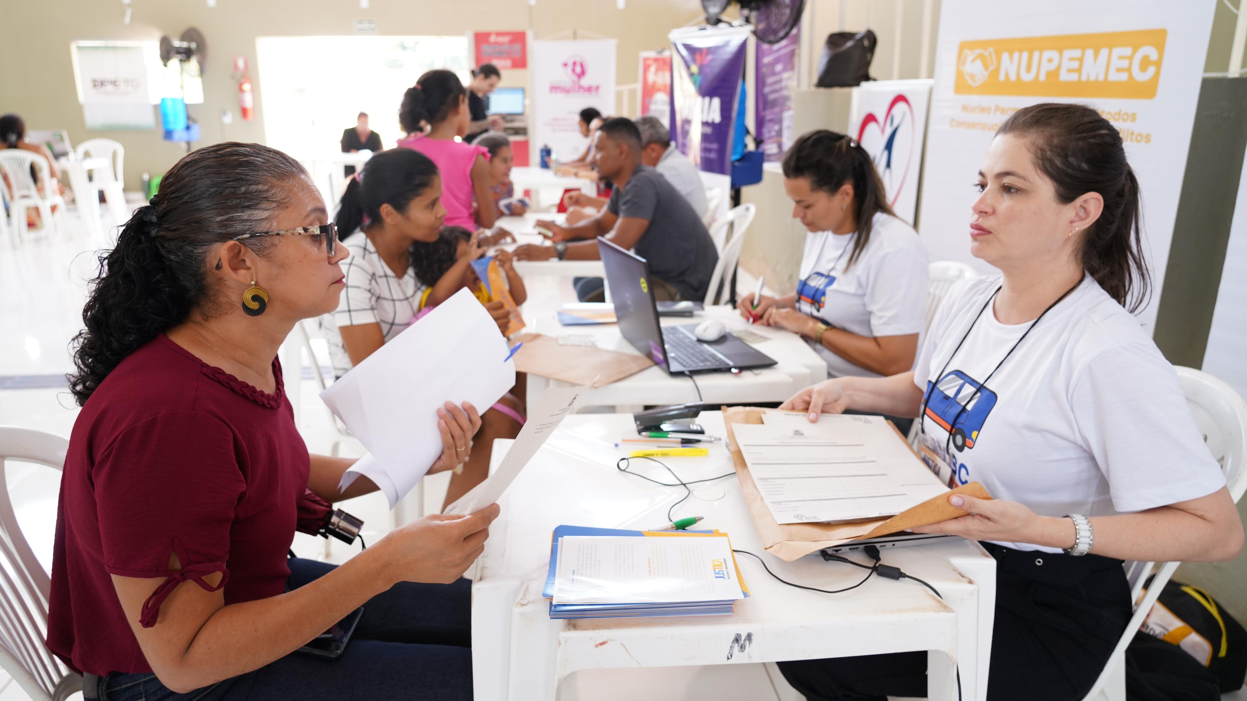 texto de audiodescrição: Fotografia colorida que mostra em primeiro plano duas mulheres sendo atendidas por outras duas mulheres do Nupemec. A imagem mostra ao fundo outras pessoas sendo atendidas