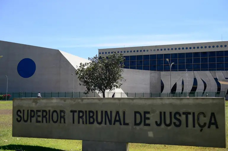fotografia da fachada do prédio do STJ em Brasília, construção em concreto armado com vitrais na cor azul, jardim e letreiro informativo escrito superior tribunal de justiça
