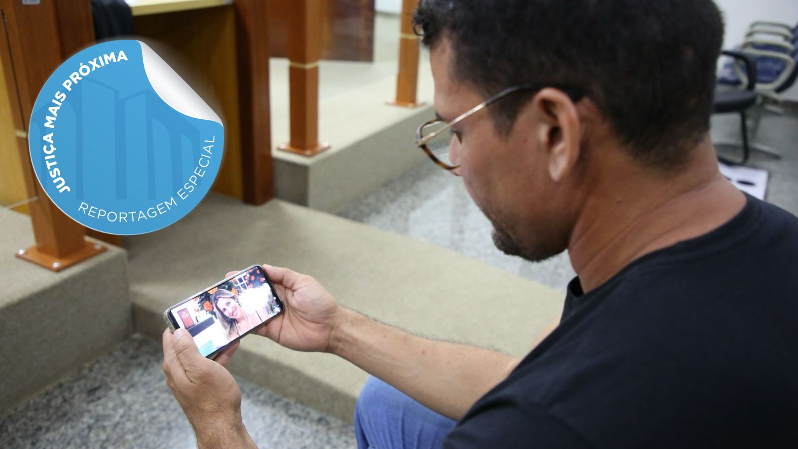 Fotografia colorida que mostra um homem jovem, usando óculos, segurando um celular com a foto da sua mãe 