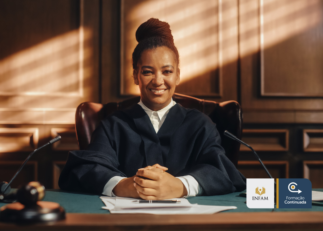 Fotografia colorida (marrom, preto, azul, branco e verde marinho) onde uma juíza negra está sentada em uma cadeira almofadada, sorrindo, de cabelos trançados e presos e ao fundo uma parede de madeira escura 