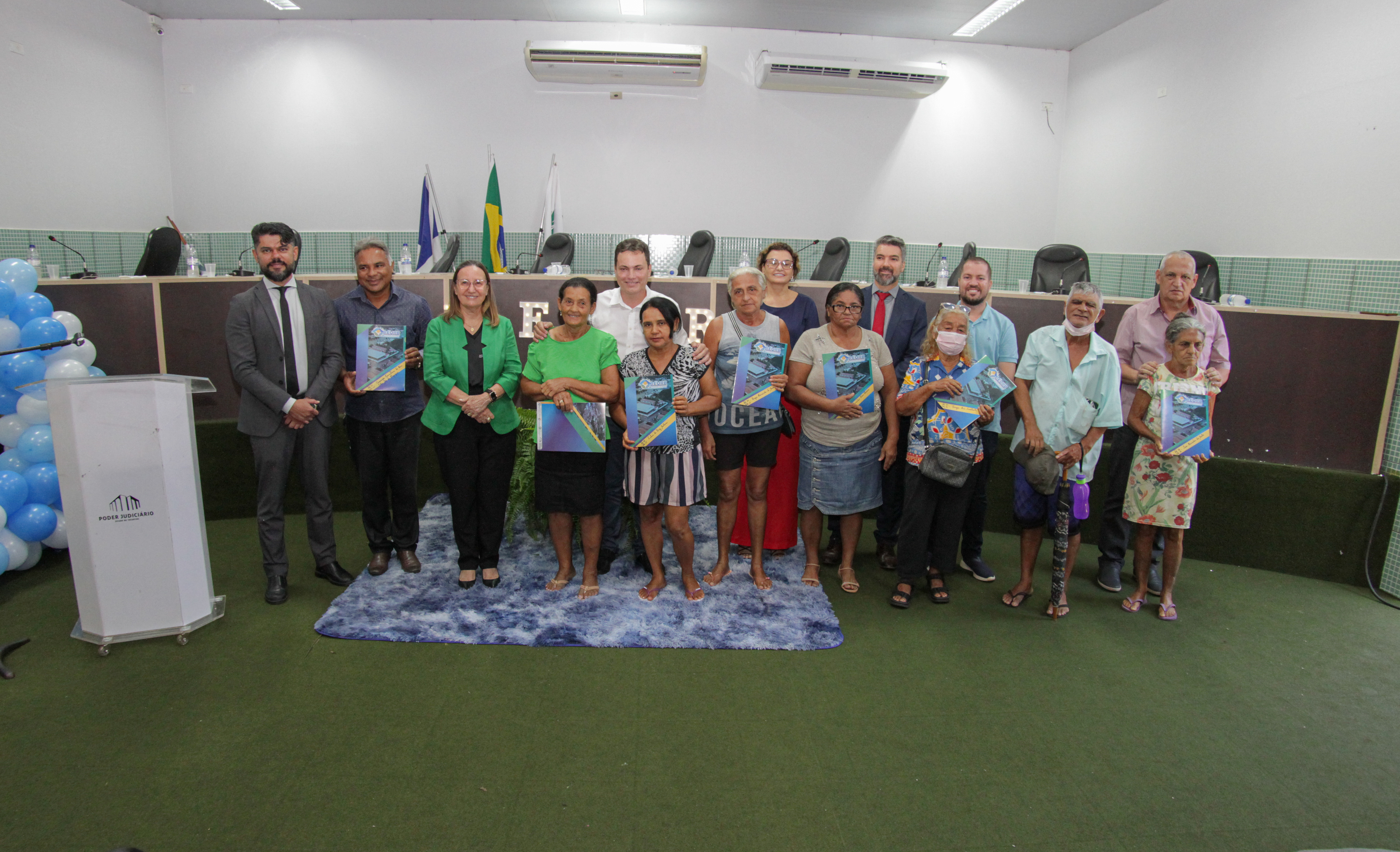 Fotografia colorida onde um grupo de moradores da cidade de Pequizeiro seguram documentos de regularização fundiária enquanto posam para foto. Ao fundo está um longo palanque escuro e ao lado um pequeno palanque branco 