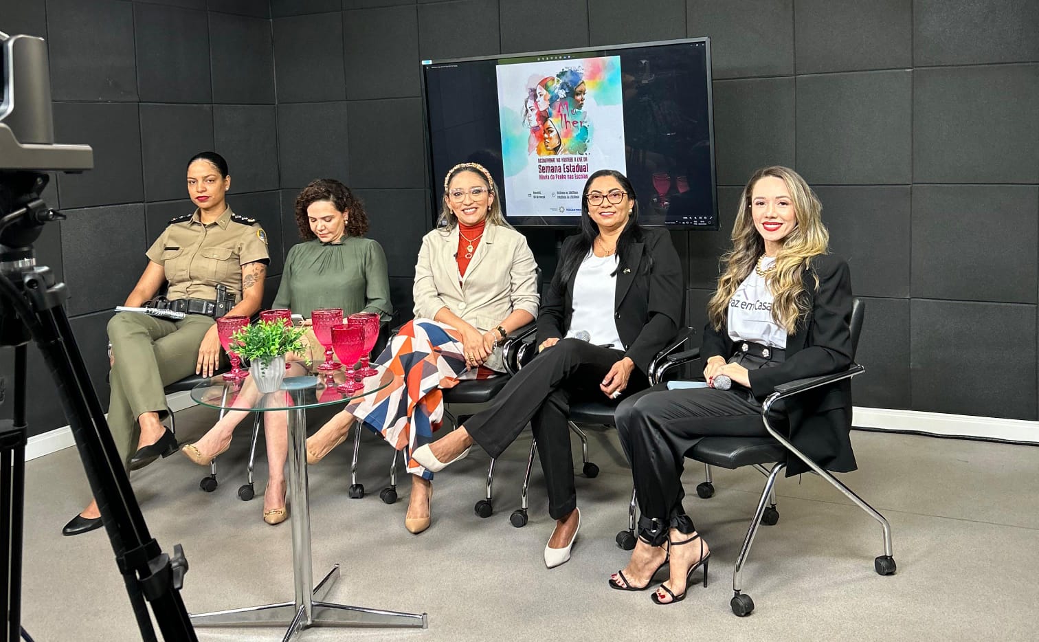 Fotografia colorida onde cinco mulheres (professora Márcia Cristina Mota Brasileiro e contou com a presença da advogada Mayana Araújo, representando a Coordenadoria Estadual da Mulher em Situação de Violência Doméstica e Familiar (Cevid/TJTO); a professora Berenice de Fátima Barbosa Castro Freitas, titular da Secretaria da Mulher; a policial militar major Flávia Roberta Pereira de Oliveira; e a promotora de justiça Munique Teixeira Vaz, do Ministério Público.) sentadas uma do lado da outra em cadeiras pretas, com uma câmera em sua frente, uma parede preta de fundo e uma televisão ativa, também de fundo.