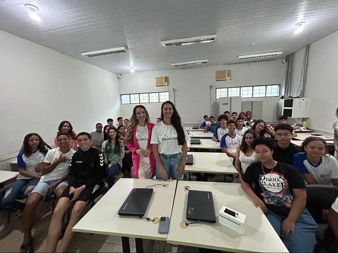 Fotografia colorida onde duas mulheres, uma de camisa branca, loira com blaze rosa, e a outra de calça, cabelos pretos e camisa branca, a camisa das duas estão com os dizeres "Justiça pela Paz em Casa", estão rodeadas de alunos de ensino médio do lado esquerdo e direito, e com mesas em suas frente e costas