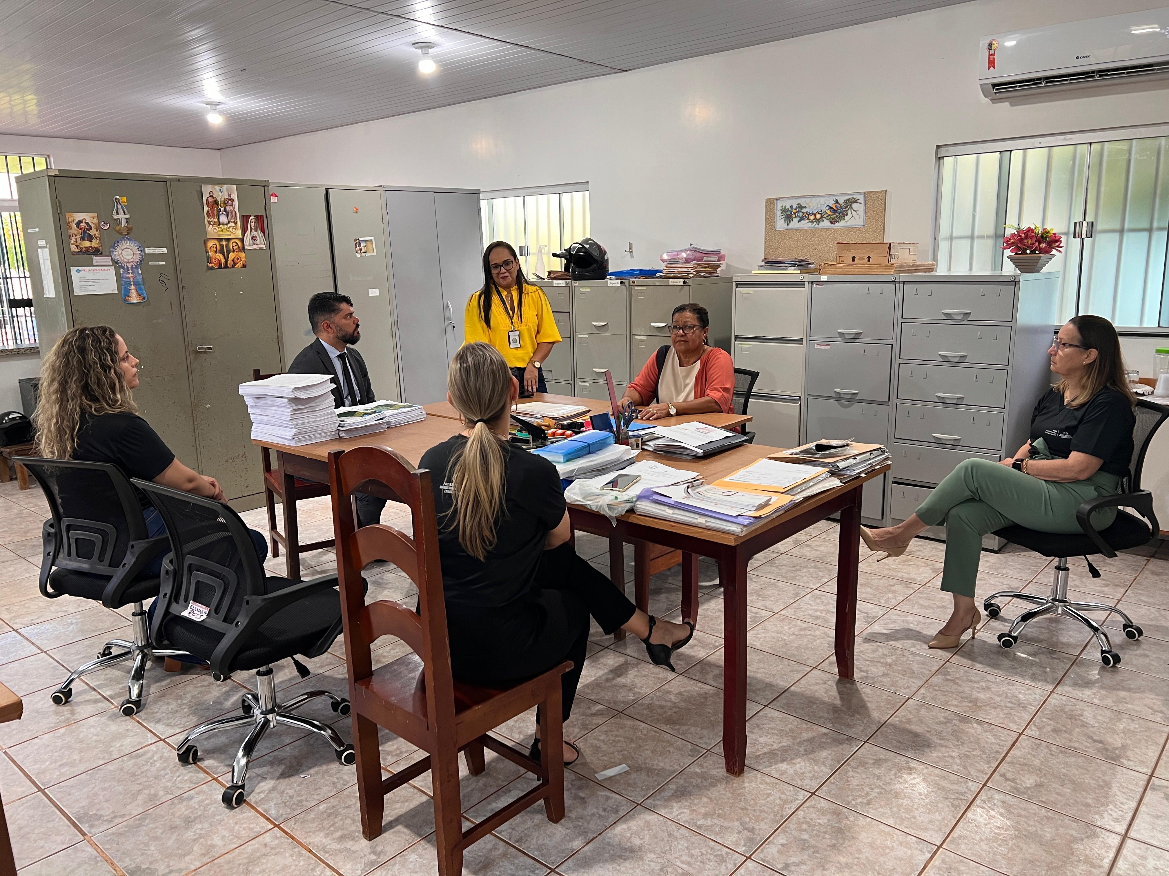 Fotografia colorida onde um grupo de cinco mulheres e um homem estão sentados em volta de uma mesa cheia de papeis conversando, em uma sala com vários armários de ferro em volta deles 