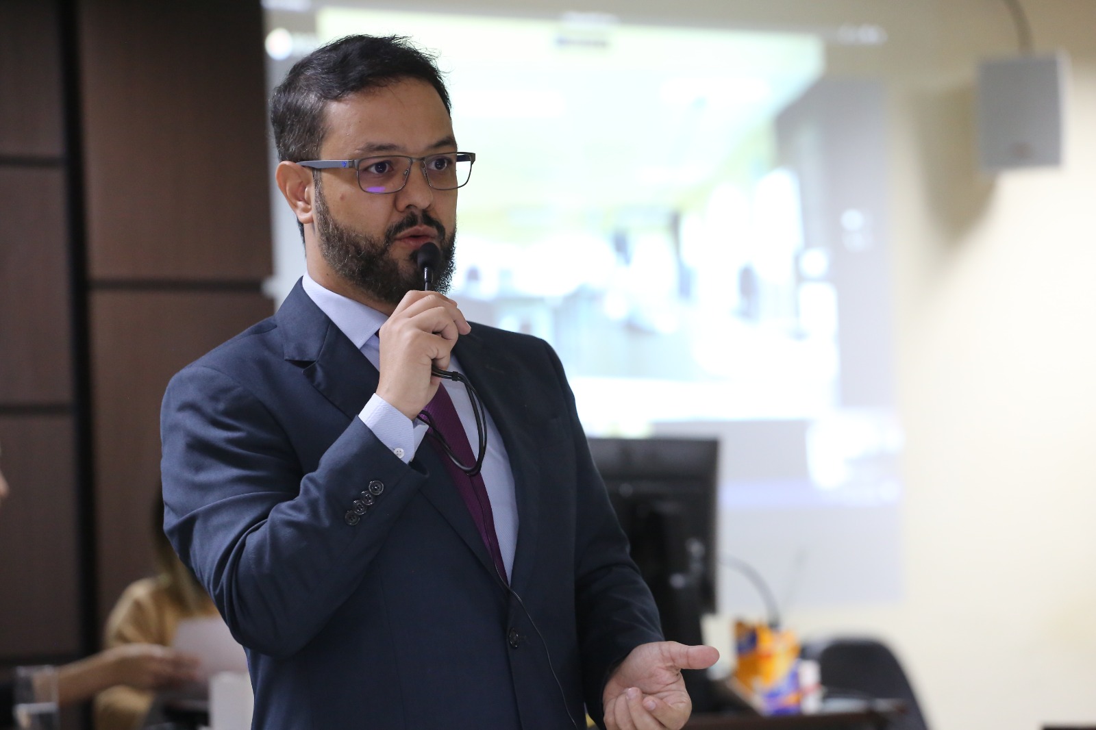 Fotografia colorida de homem pardo, barbudo,de terno e óculos,segurando um microfone 