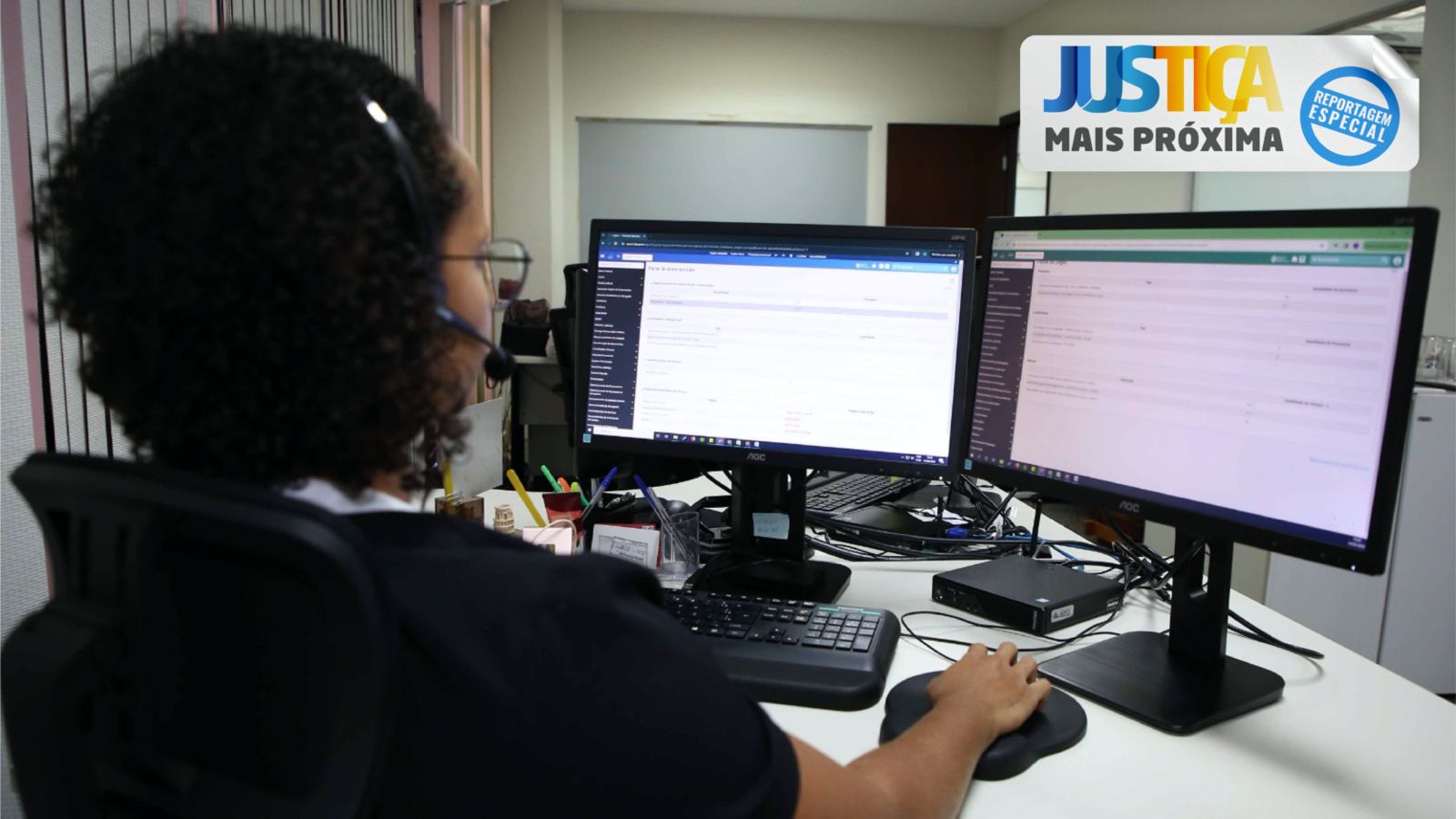 Mulher de cabelos pretos e curtos abrindo o sistema Eproc no computados à sua frete, ela está sentada em uma mesa e aparecem dois monitores; na parte superior à direita, a logo da Justiça Mais Próxima 