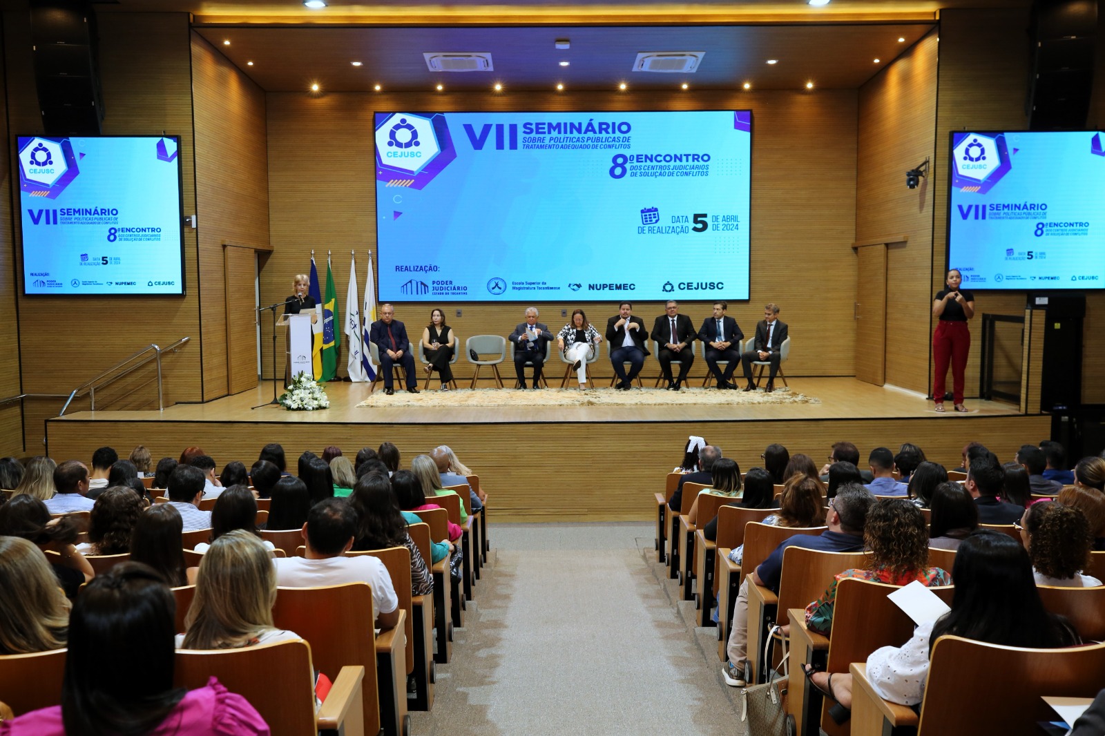 Imagem de pessoas sentadas em auditório assistindo mulher branca de cabelos loiros falando ao púlpito, enquanto outras pessoas estão sentadas ao seu lado. Ao fundo, na parede revestida de madeira, três telões projetam imagem do evento em tons azul, Seminário sobre resolução de conflitos