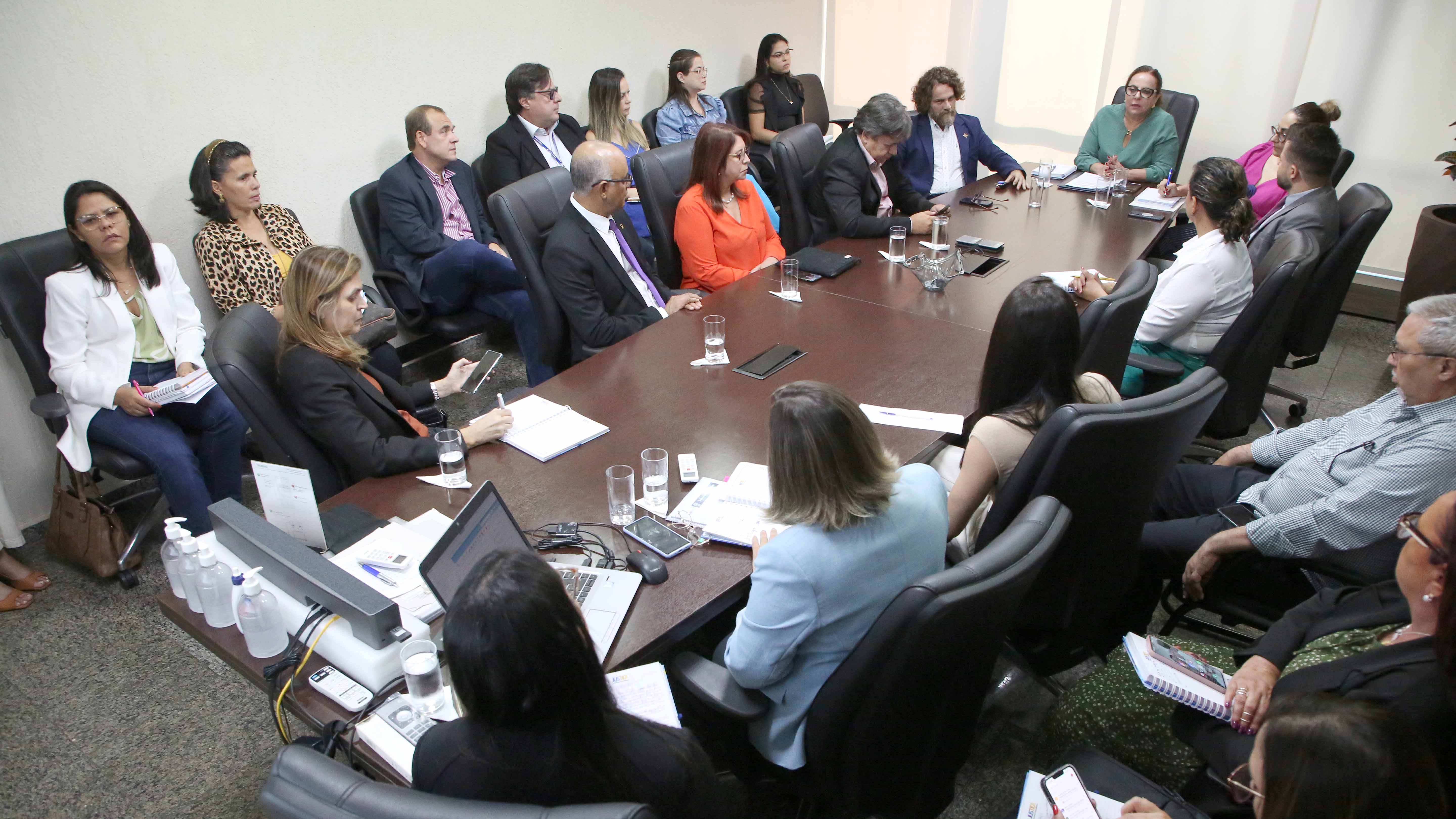 Imagem de homens e mulheres reunidos ao redor de uma mesa de madeira em tom escuro, em uma sala branca com persiana clara