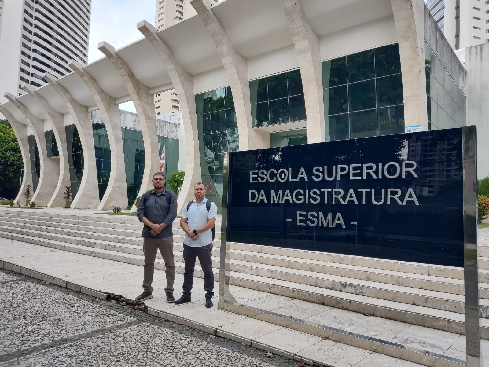 Dois homens em pé em uma pequena escada em frente a um prédio branco com películas de vidro verde, ao lado de uma placa da Escola Superior da Magistratura