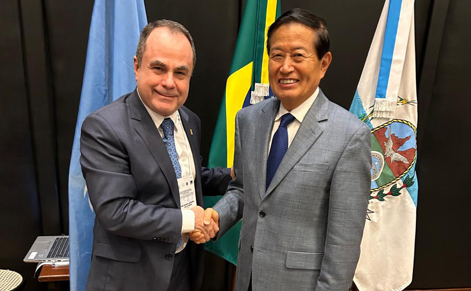 Imagem colorida que mostra dois homens (um oriental e outro brasileiro) num aperto de mãos. Eles vestem terno e  gravata e estão em uma sala fechada com três bandeiras atrás