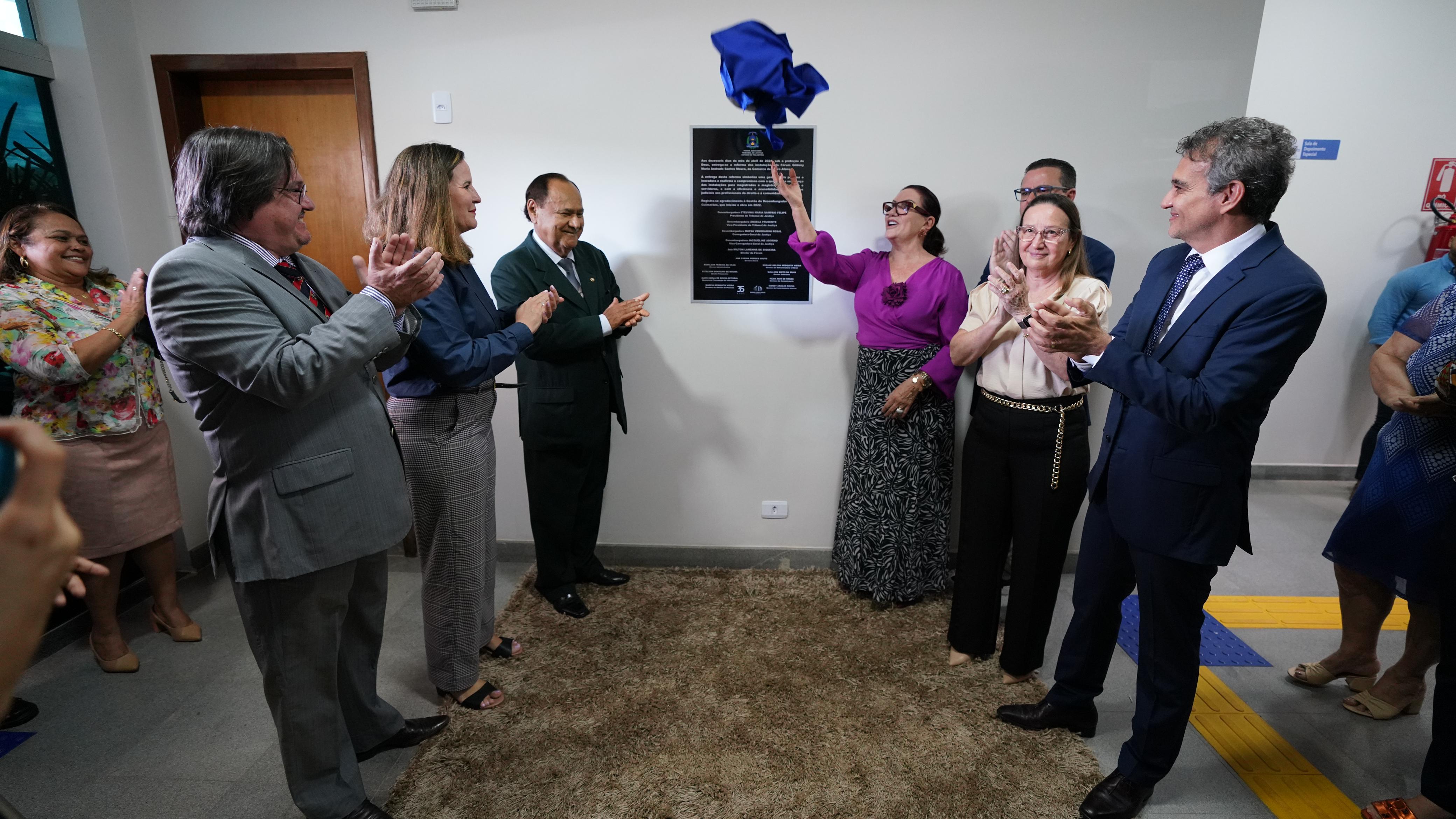 Homens e mulheres em pé, sobre um tapete marrom, em frente a uma placa instalada numa parede branca, durante o seu descerramento na inauguração da reforma do Fórum de Pedro Afonso. O tecido azul da placa é retirado pela mulher de saia preta longa e blusa de manga comprida rosa fúcsia, presidente do TJ, desembargadora Etelvina, que usa óculos de grau e cabelos presos