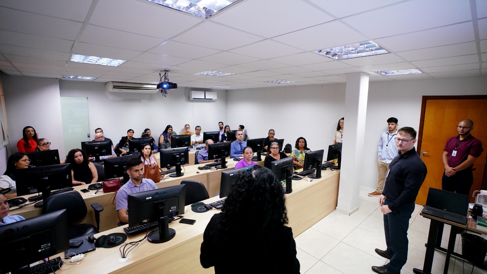 Homens e mulheres sentados em um laboratório de informática durante oficina ministrada por professores homens e mulheresevemente inclinado orientando aluna sentada, loira de cabelo em coque, e camisa azul