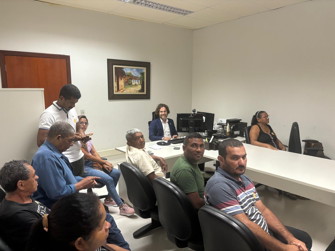 Imagem colorida que mostra homens e mulheres reunidos numa sala, em volta de uma mesa. No centro da mesa está um homem jovem, claro, de cabelos longos - ele usa terno e gravata
