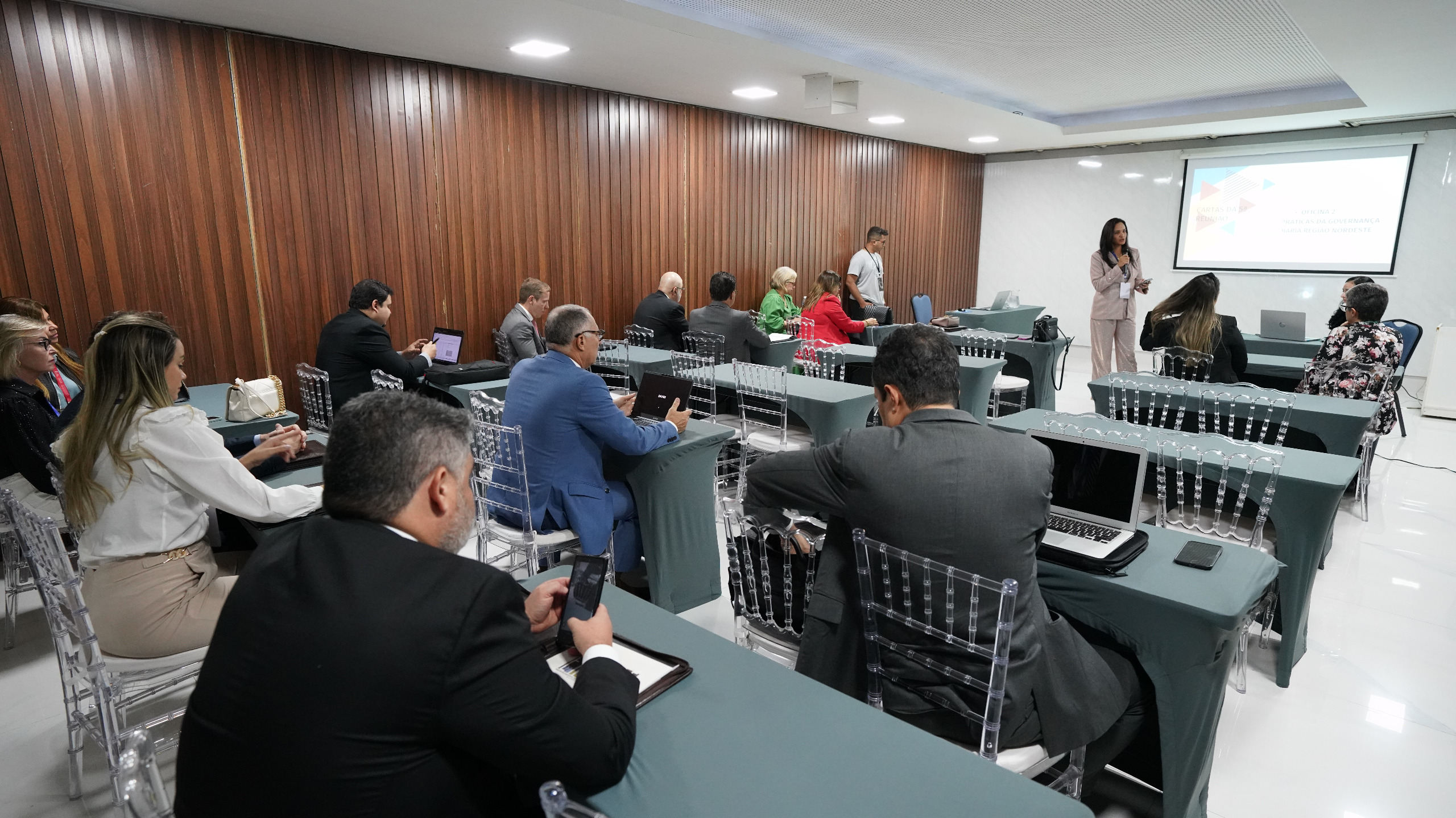 Mulher com roupa clara está à frente de um telão e fala para várias pessoas que estão sentadas