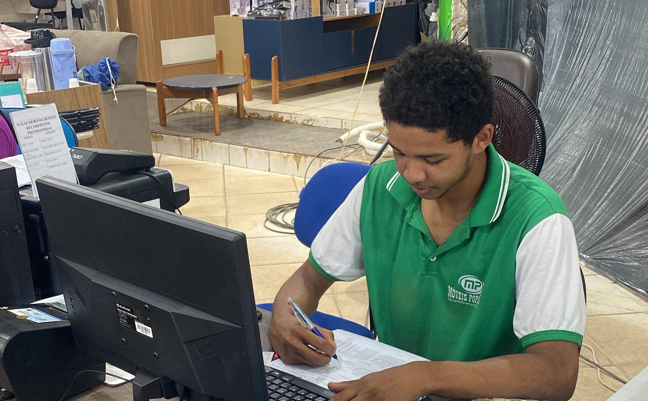 Fotografia colorida que mostra um jovem rapaz negro usando uma roupa verde e escrevendo no caderno
