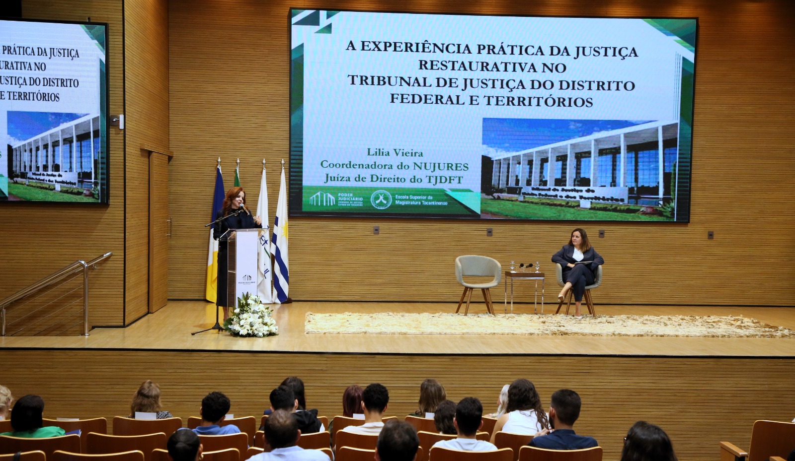 Fotografia colorida onde uma mulher fala em um palanque de acrílico, em cima de um palco de madeira, enquanto outra está sentada em uma cadeira almofadada, ao lado de outra cadeira igual, com uma mesa entre elas, e no fundo um slide passando na tela 