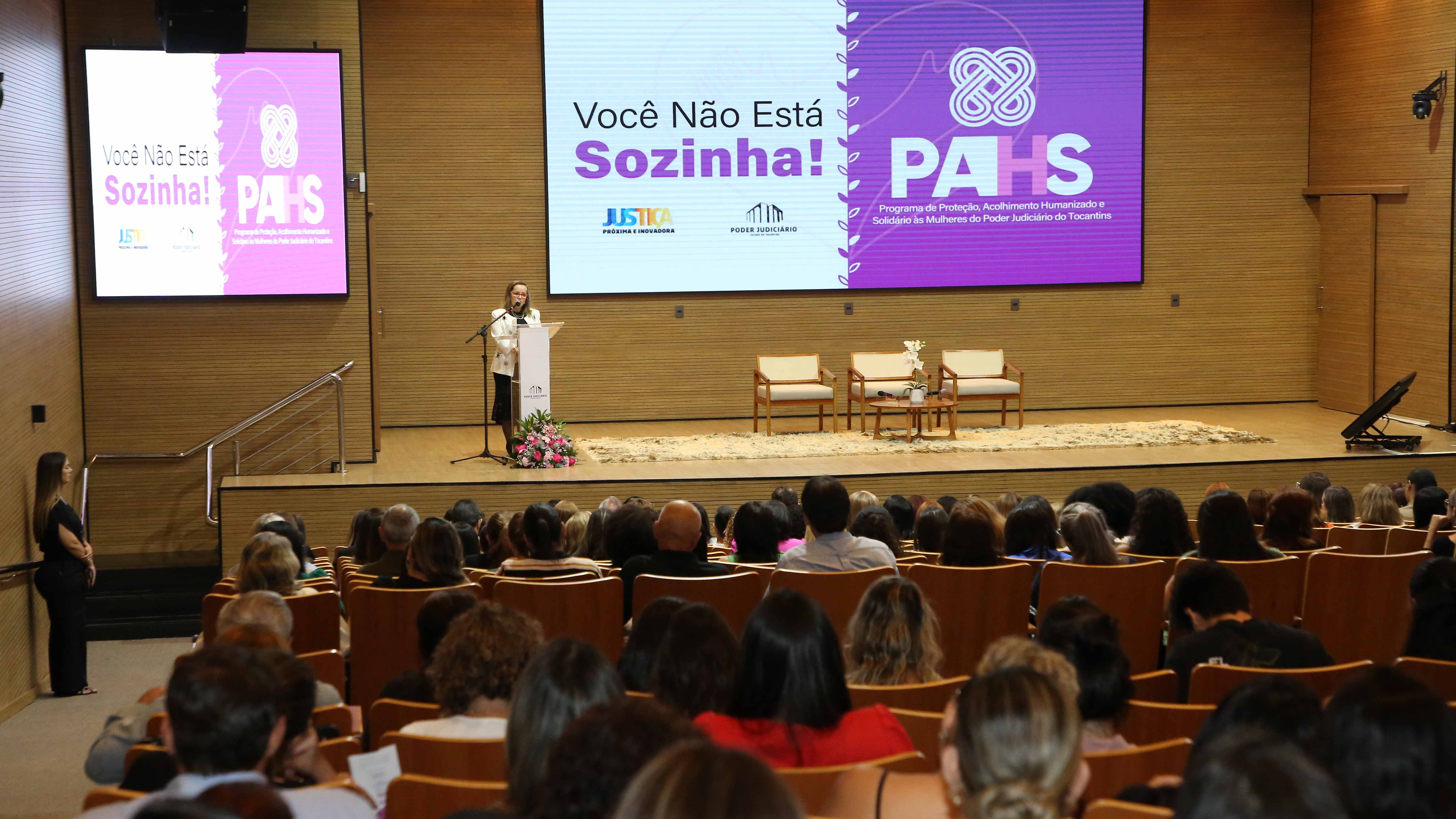 Imagem aberta do audiório, com várias pessoas sentadas e a juíza Cirlene ao fundo, no palco, falando ao microfone 