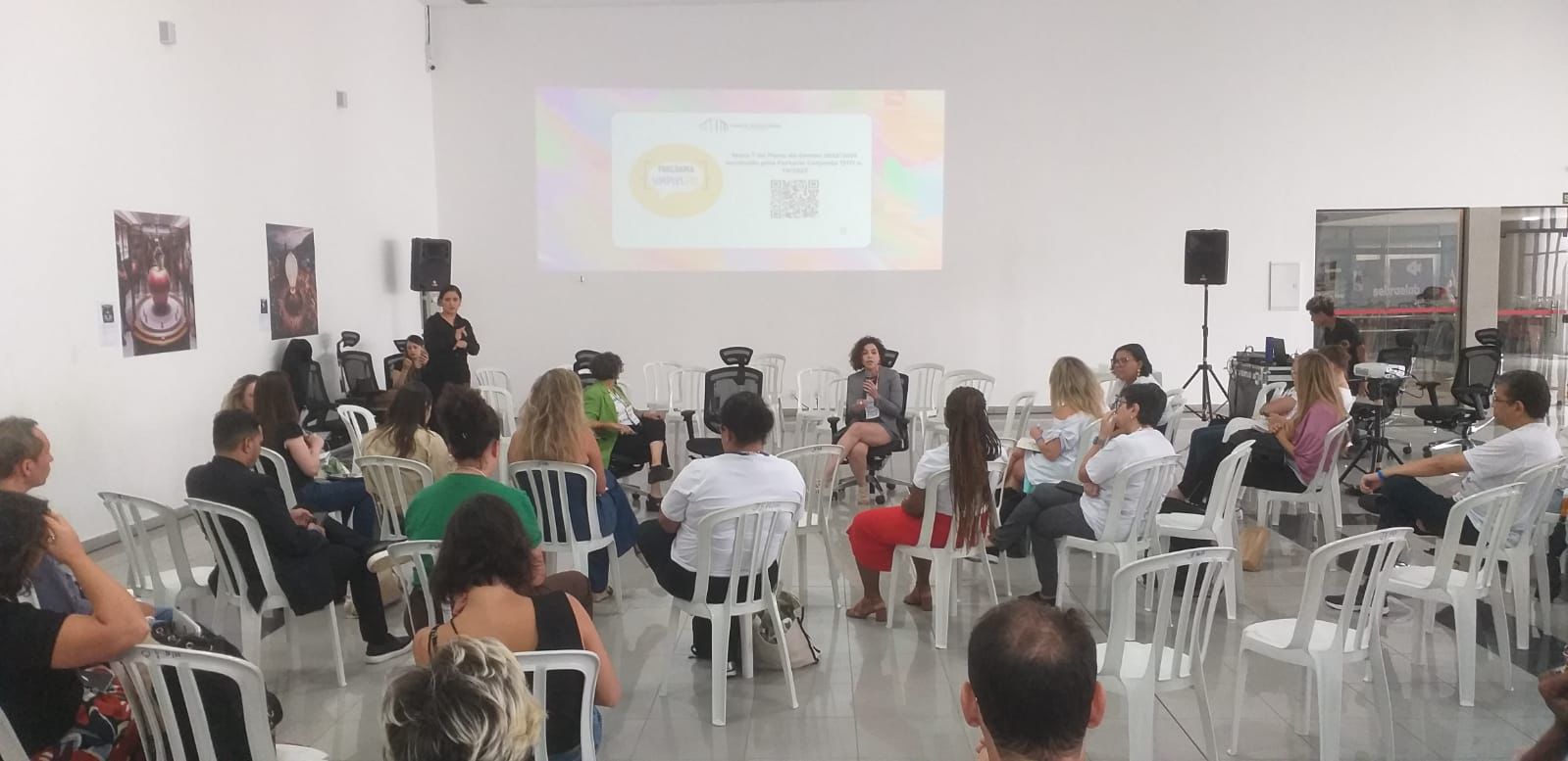 várias pessoas sentadas em cadeiras brancas assistindo a palestra da coordenadora Roberta Maciel, que está de frente; ao fundo um telãoprojetando imagens