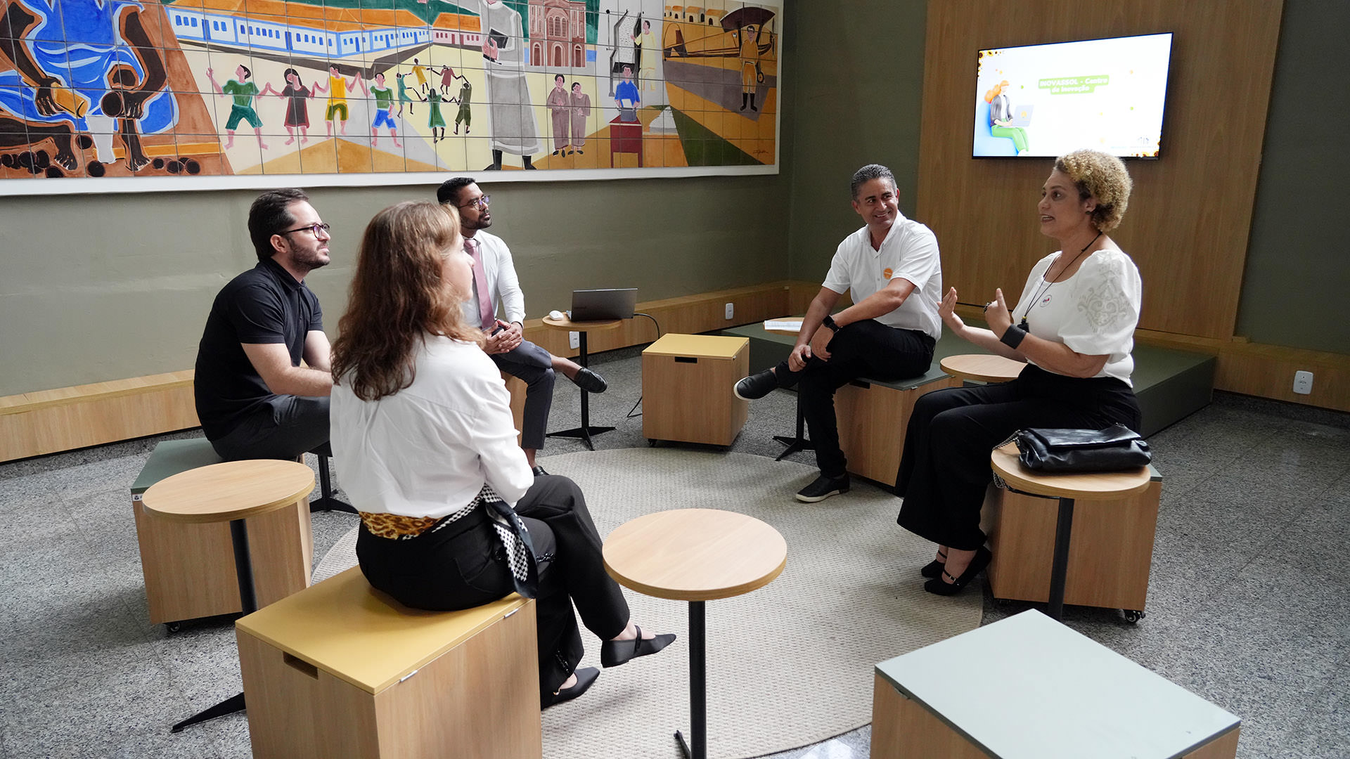 Imagem mostra um grupo de pessoas sentadas em um círculo, em frente a uma parede com uma grande pintura. As pessoas estão em reunião, sentadas em circulo..