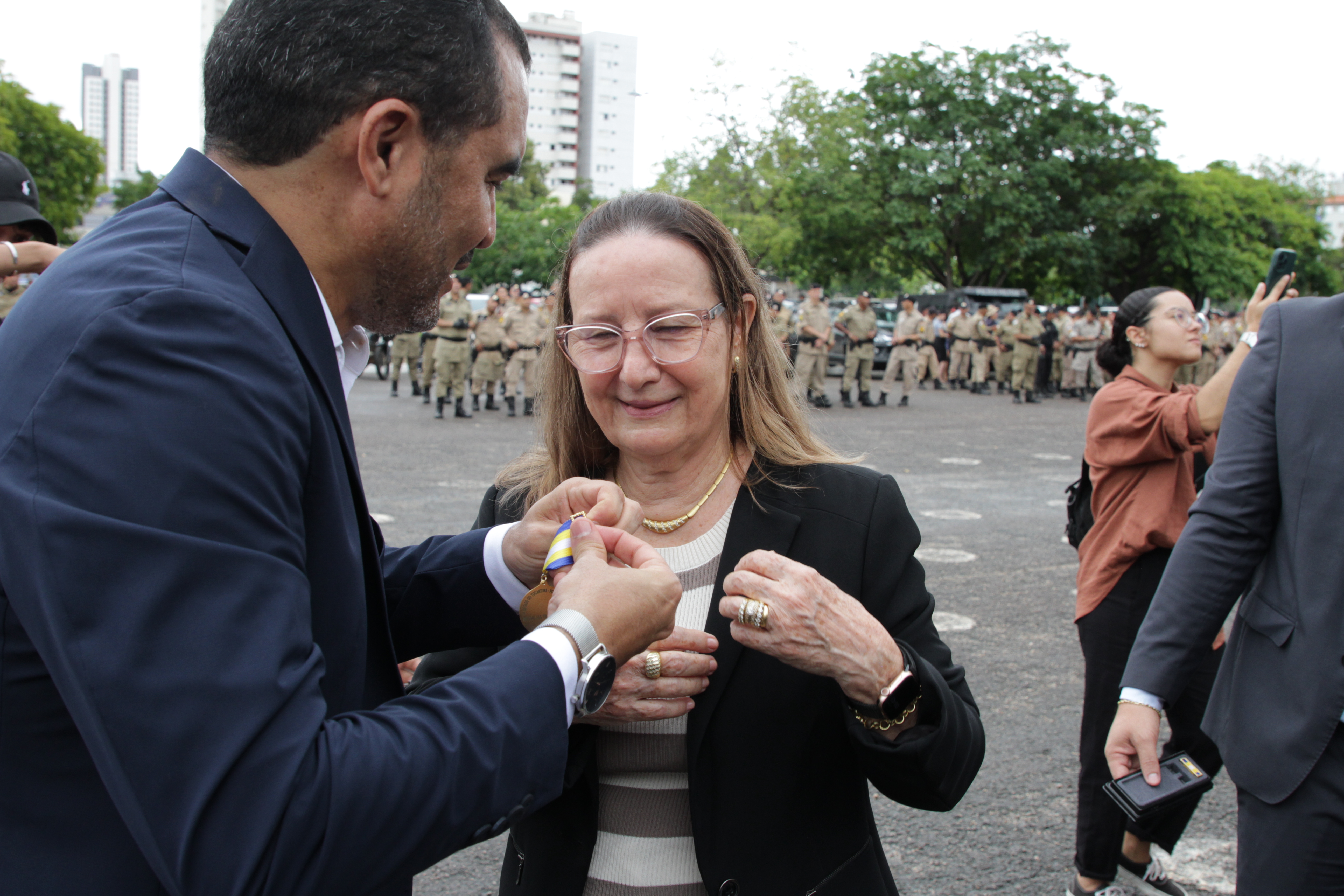 Fotografia colorida onde a corregedora-geral desembargadora Maysa Vendramini Rosal das mãos do governador  Wanderlei Barbosa. Ela está de blazer preto, com uma camisa listrada das cores branco e bege, cabelo solto castanho claro e óculos, e o governador está de terno e camisa branca de botão. Ao fundo, policiais estão em fileiras 