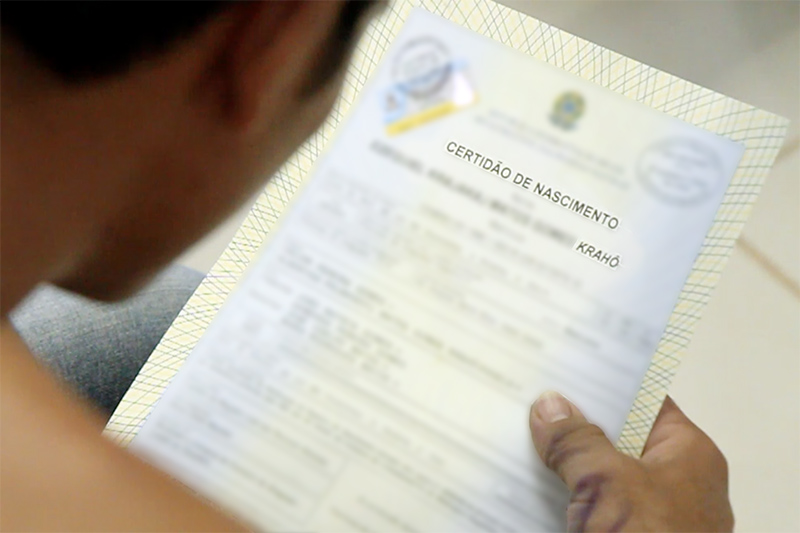 imagem de detalhe da cabeça de um indígena com uma certidão de nascimento (em destaque) na mão