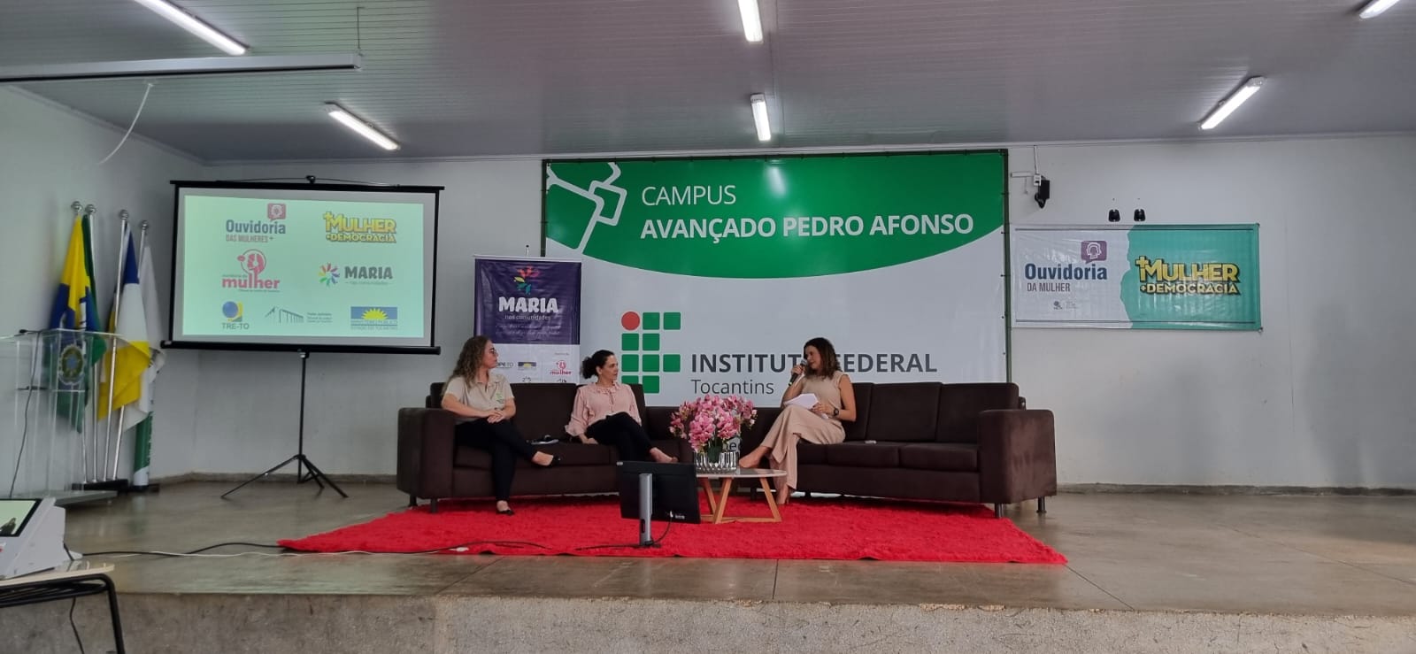 Fotografia colorida onde três mulheres estão sentadas em dois sofás  marrons, com um tapete vermelho grande no chão, uma televisão ao lado e ao fundo um grande banner do câmpus avançado do IFTO de Pedro Afonso 