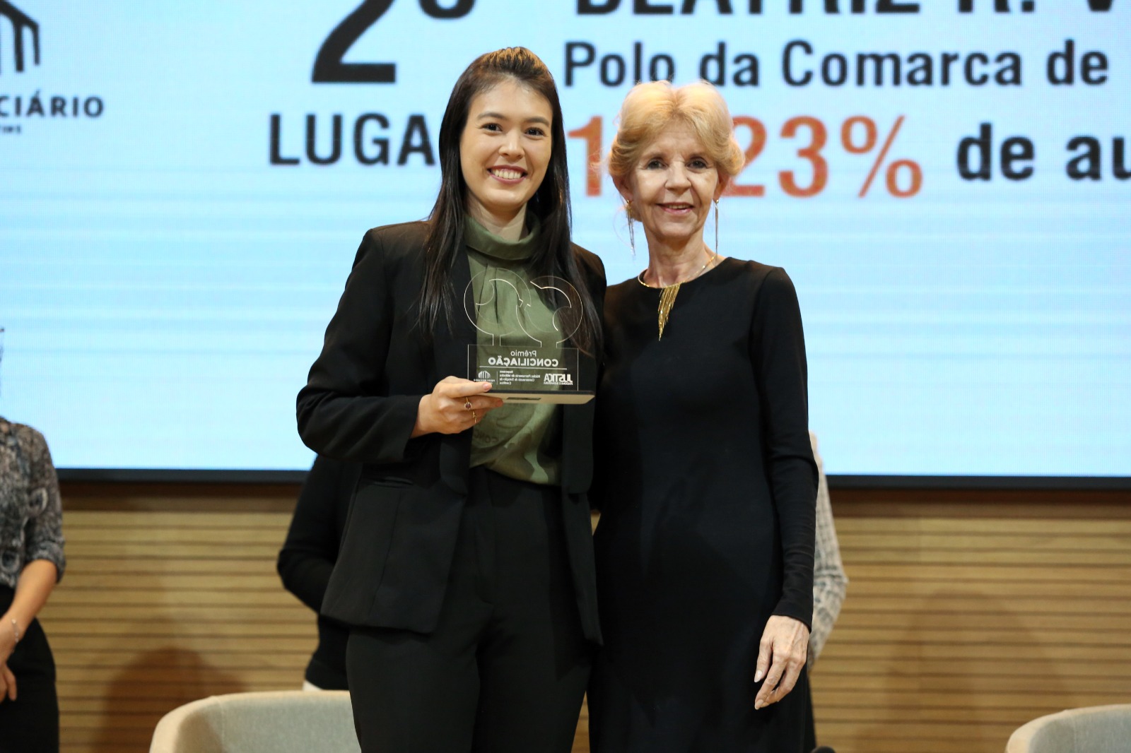 juíza Silvana Parfieniuk, uma mulher de cabelos louros e roupa preta, com a primeira colocada, uma jovem de cabelos lisos pretos e roupa escura segurando o trofeu