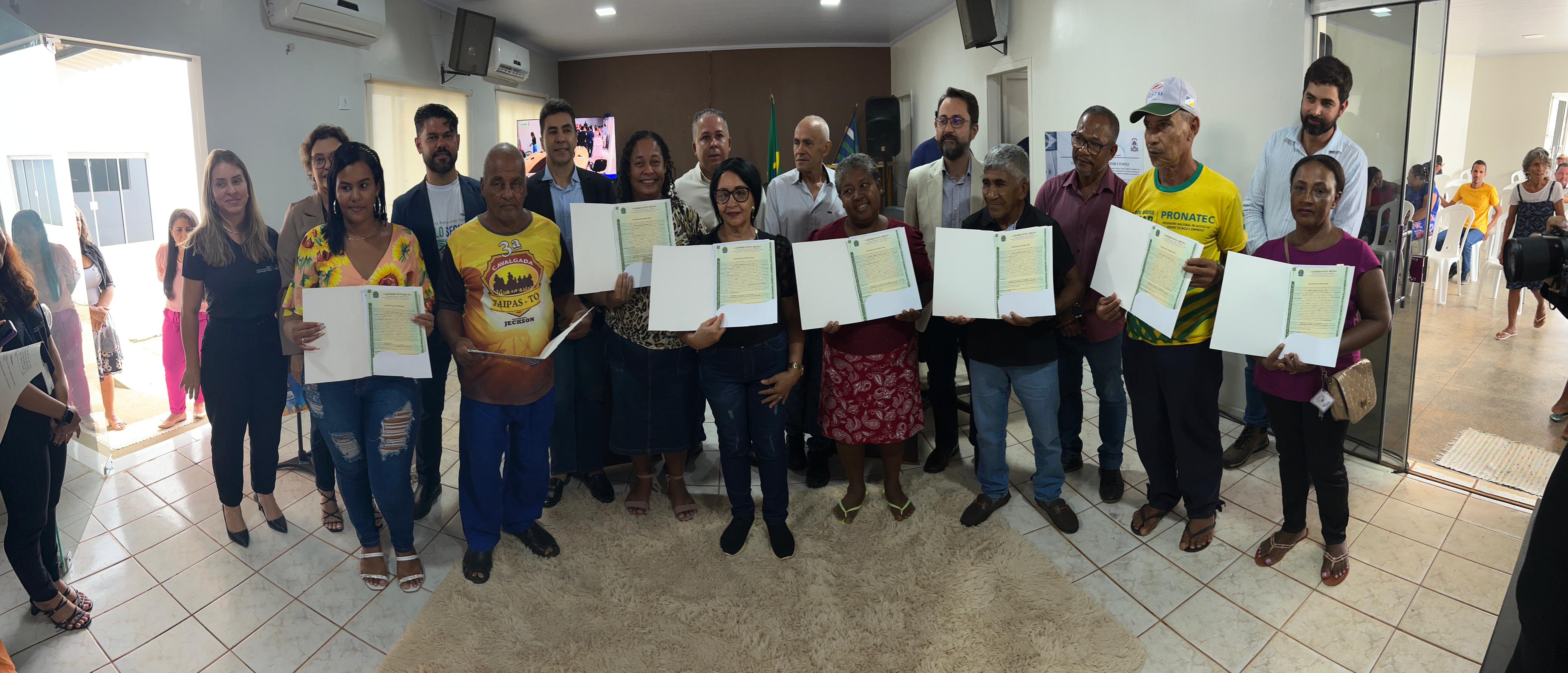 Fotografia colorida onde um grupo de dezessete pessoas estão em pé, uma do lado da outra, segurando documentos de regularização fundiária  