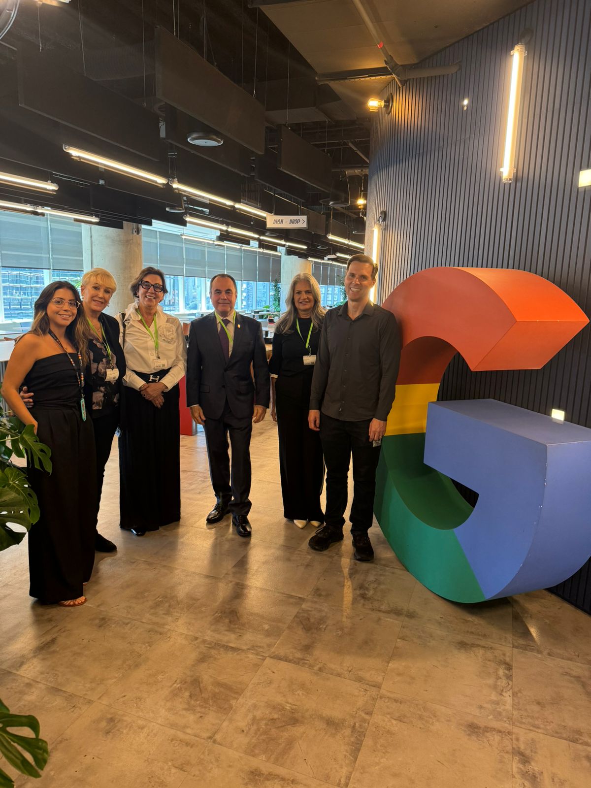 Fotografia colorida onde seis pessoas, dois homens e quatro mulheres, estão posando sorrindo para foto lado a lado em um escritório com mesas e um teto com -placas retangulares ao fundo, ao lado de uma grande escultura da letra G do Google