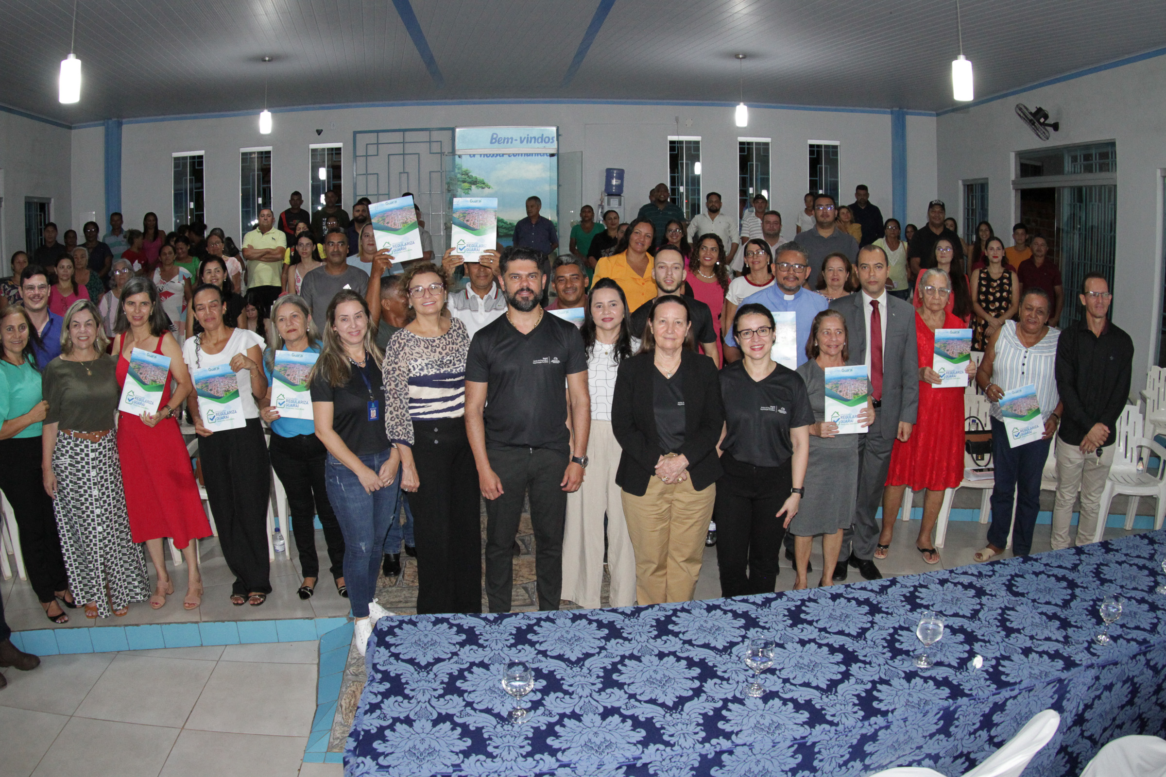 Fotografia colorida onde várias pessoas estão agrupadas posando para foto. Alguns estão com documentos de regularização fundiária nas mãos, e estão dentro de uma sala branca com linhas azul claro no teto e no chão. Em sua frente, está uma mesa com uma manta azul escuro em padrões floridos 