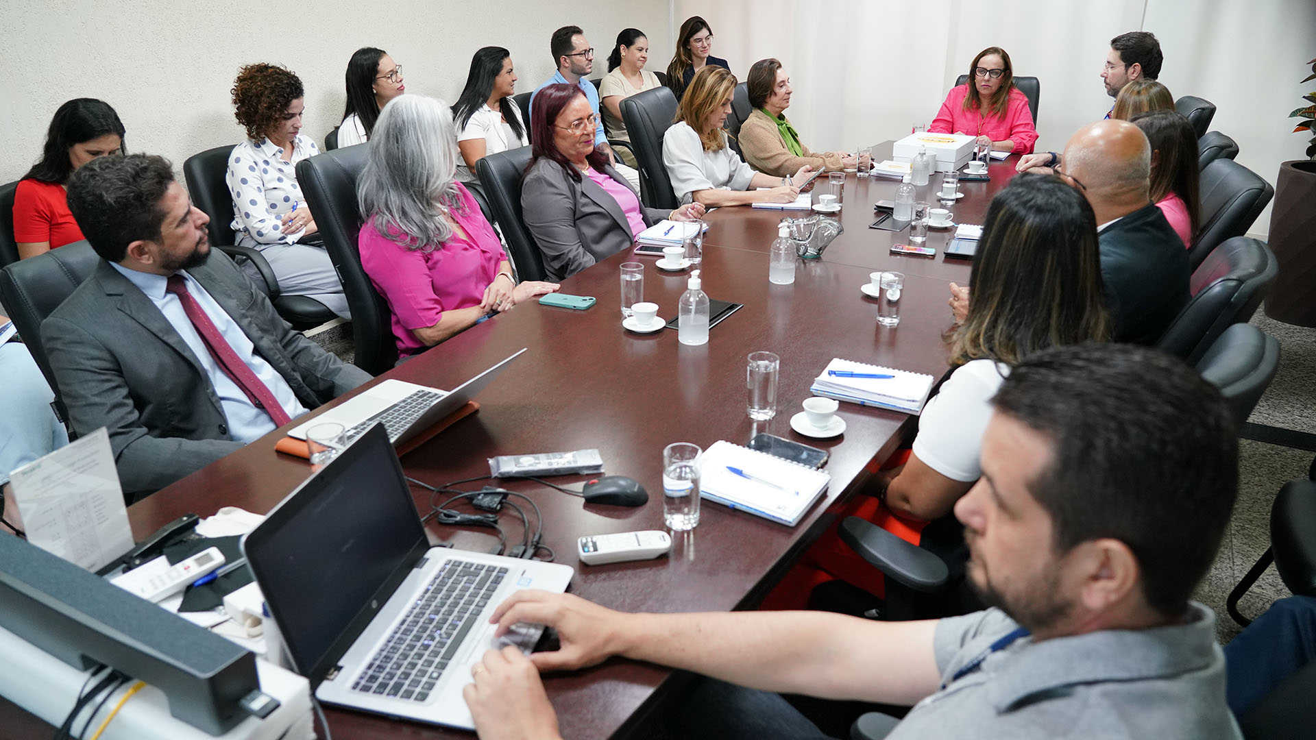 Em uma sala várias pessoas estão sentadas em cadeiras e a mesa é de madeira oval, têm copos de água nas mesas e as pessoas estão falando