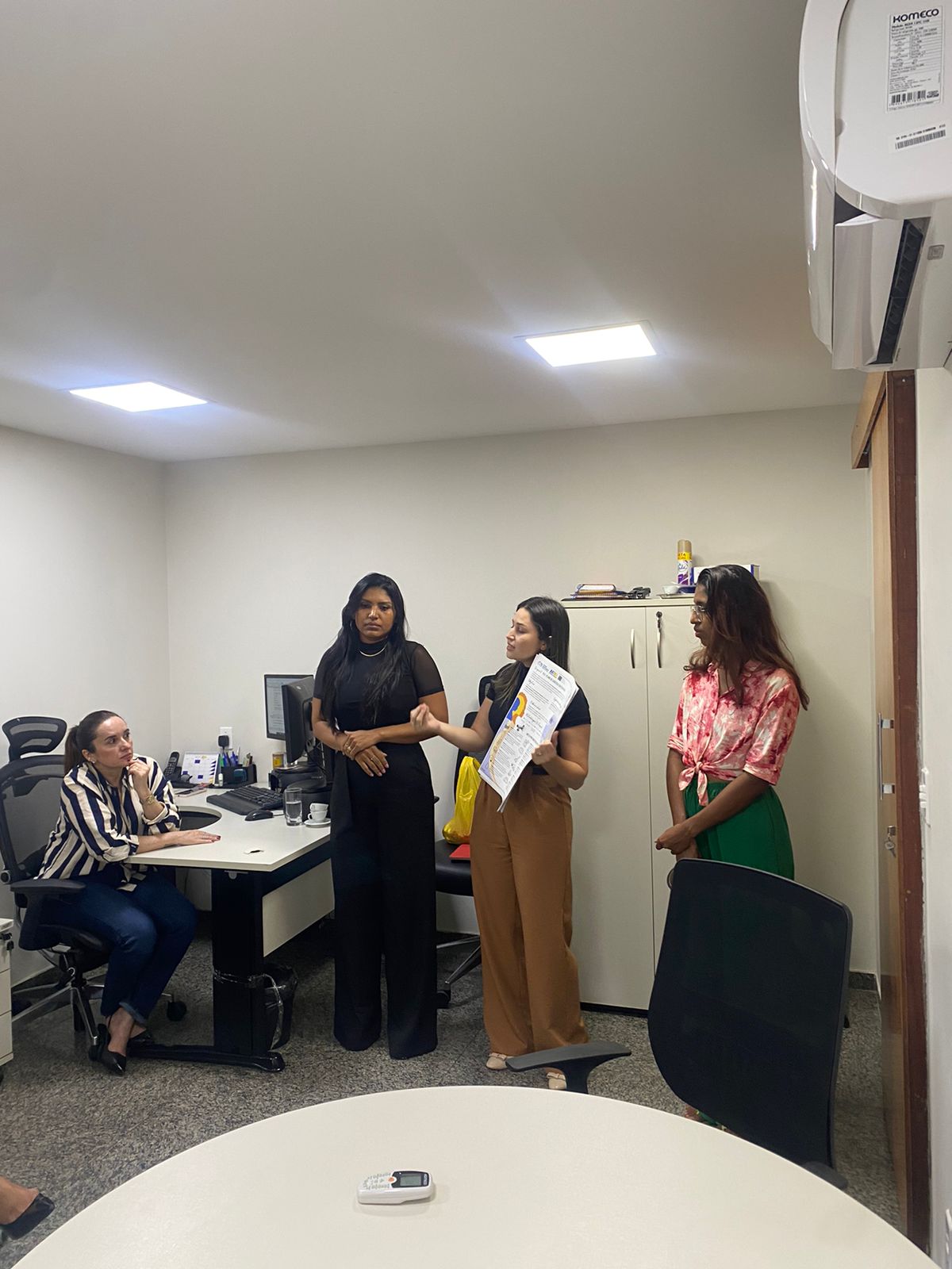 Três mulheres em pé, falando para uma pessoa. Elas seguram um plano impresso na mão