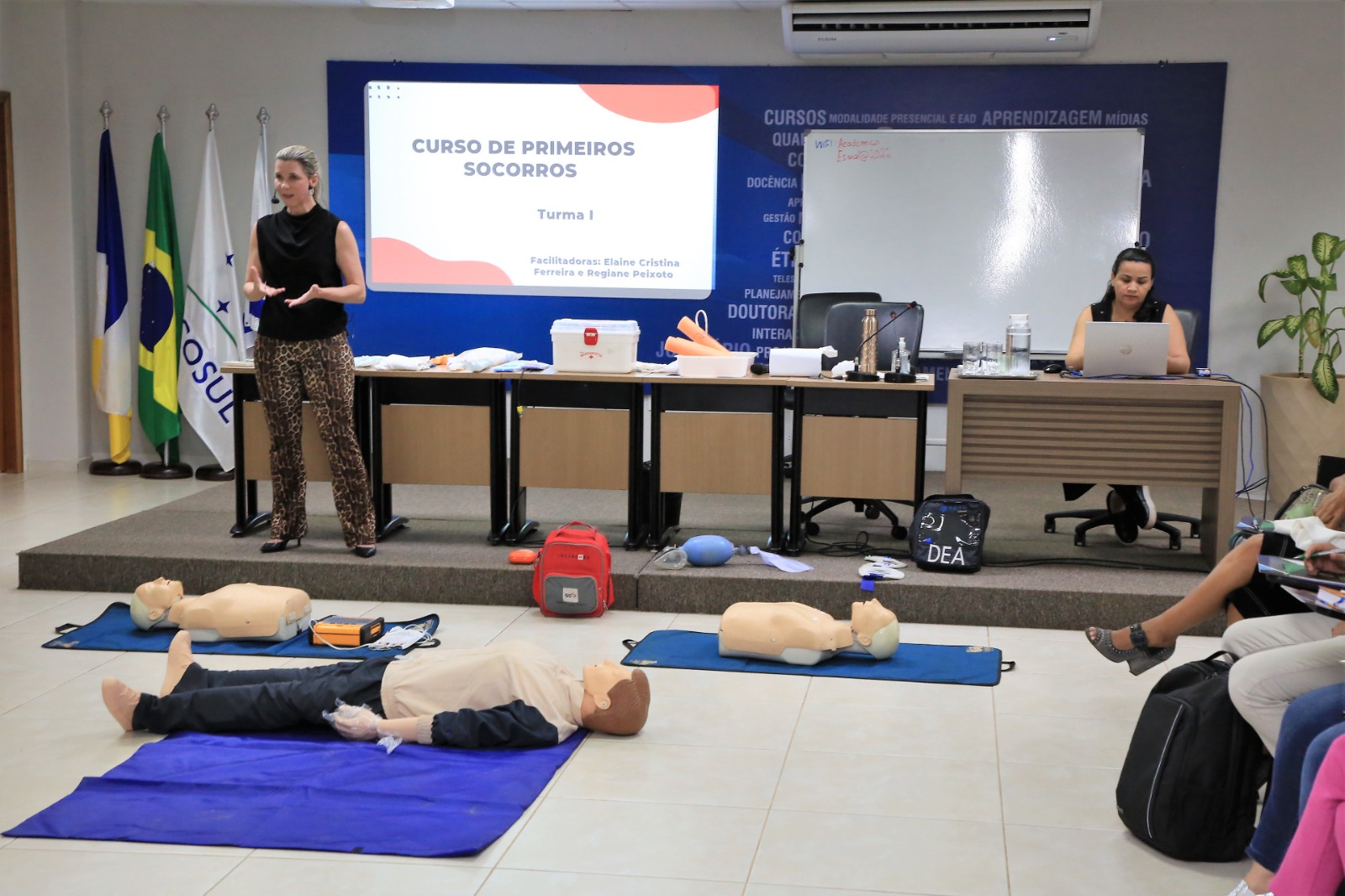 Mulher branca de cabelos loiros amarrados veste blusa preta sem manga e calça com estampa de onça e sapato preto. Ela tá em pé em frente a uma mesa de madeira dando curso de primeiros socorros, ao fundo uma tela está em cima de um fundo azul e projeta inscritos do tema. Tem quatro bandeiras no canto esquerdo e no canto direito uma moça parda de cabelos liso e soltos na altura do ombro sentada ao computador. À frente, três bonecos deitados em tatames azul no chão para simular massagem toráxica
