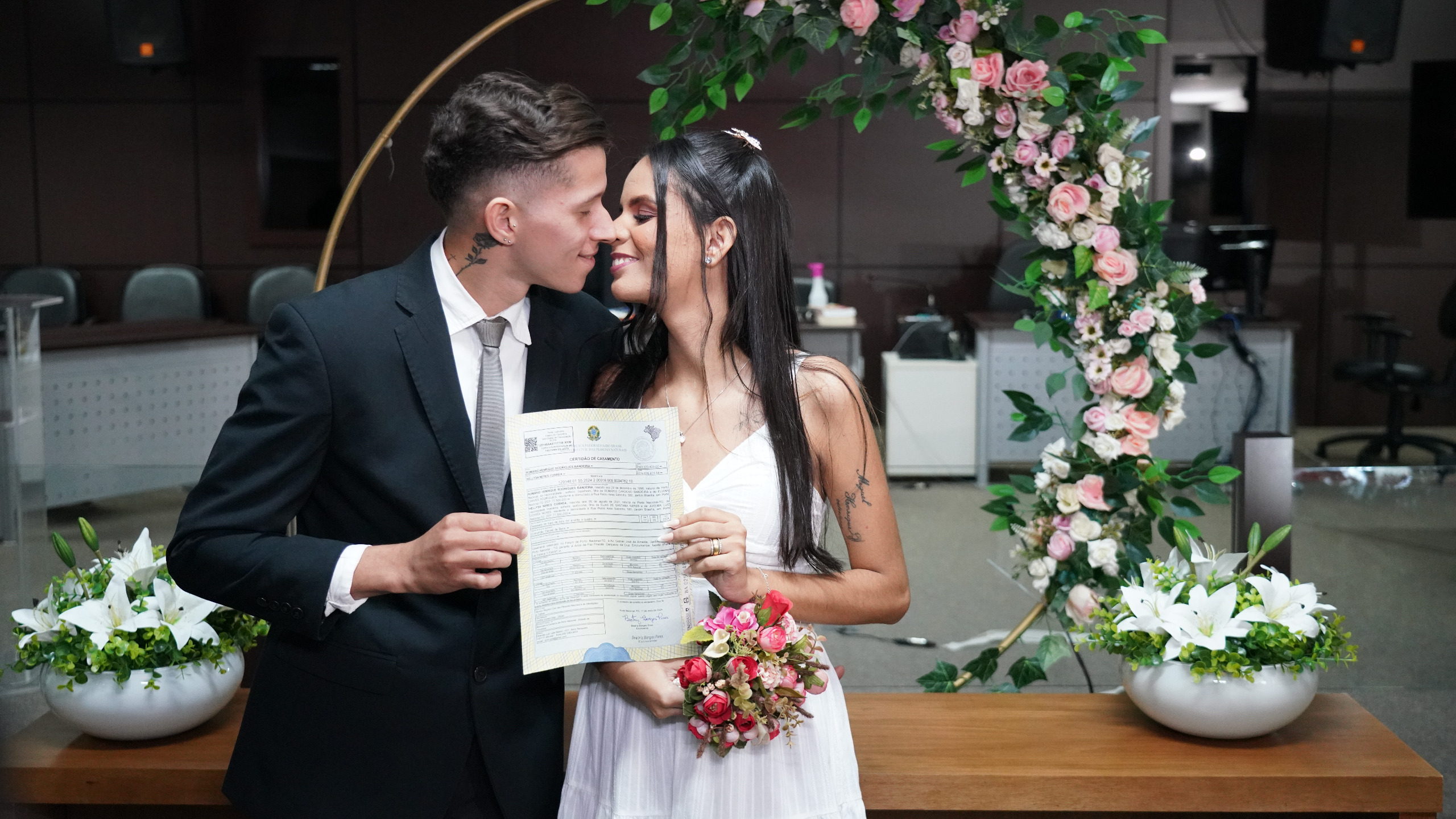 homem de terno preto e camisa branca beija mulher vestida de noiva, o casal segura o documento de certidão de casamento