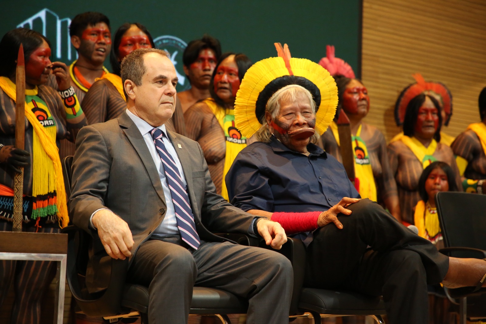 a esquerda homem branco usa terno e gravata e à direita, indígena usa vestes tradicionais atras dos dois vários indígenas usam roupas tradicionais