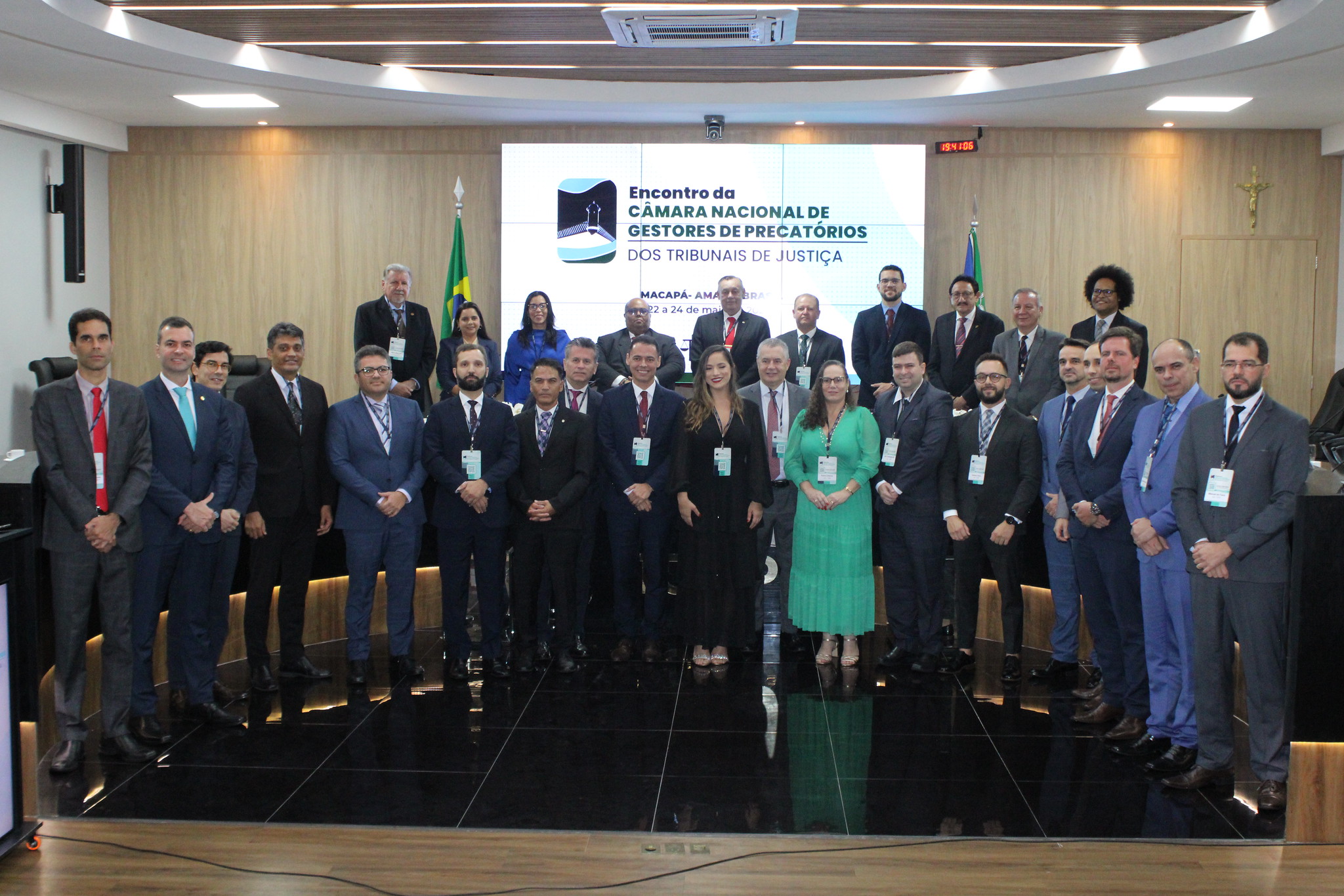 Homens e mulheres em pé em frente a um telão centralizado numa parede revestida de madeira num tom claro. O telão projeta dizeres sobre Encontro Nacional de Gestores de Precatórios dos Tribunais de Justiça do Brasil