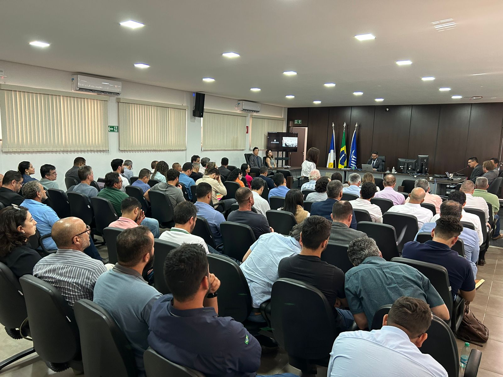 Foto mostra várias pessoas sentadas em cadeiras pretas e à frente uma mesa e uma pessoa sentada falando ao microfone