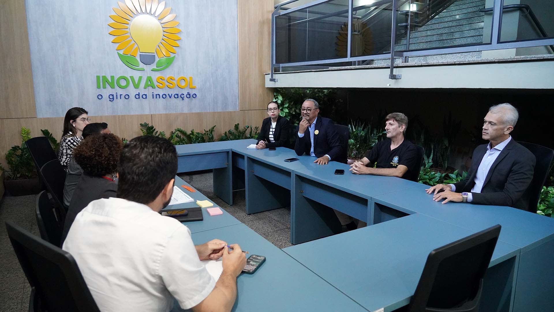 Fotografia colorida que mostra homens e mulheres sentados à mesa; ao fundo, vê-se uma imagem de um girassol escrito: Inovassol