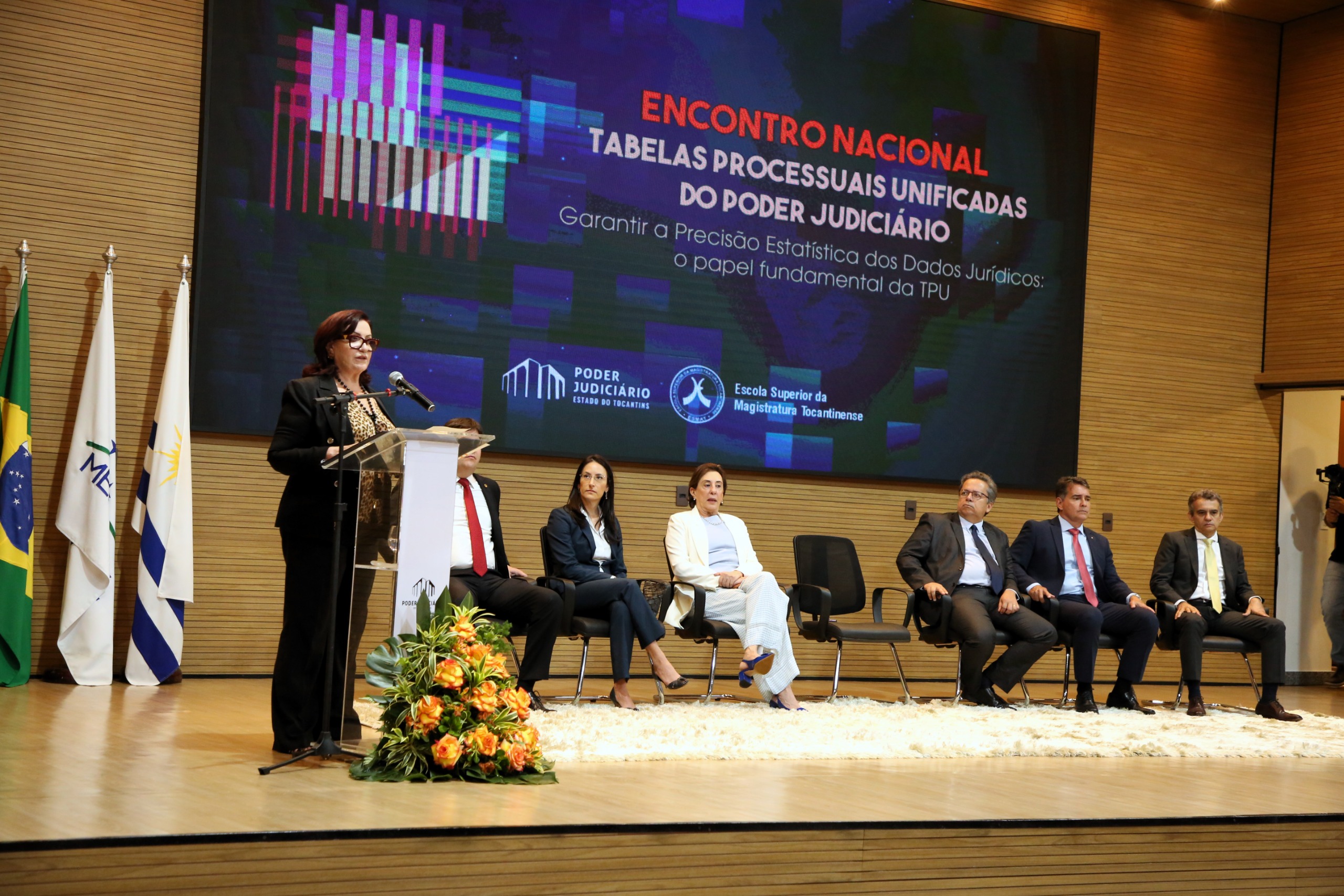 Desembargadora Etelvina Sampaio fala ao microfone durante abertura do evento, observada por demais autoridades no palco do auditório; ela usa óculos, blusa estampada e terninho escuro 