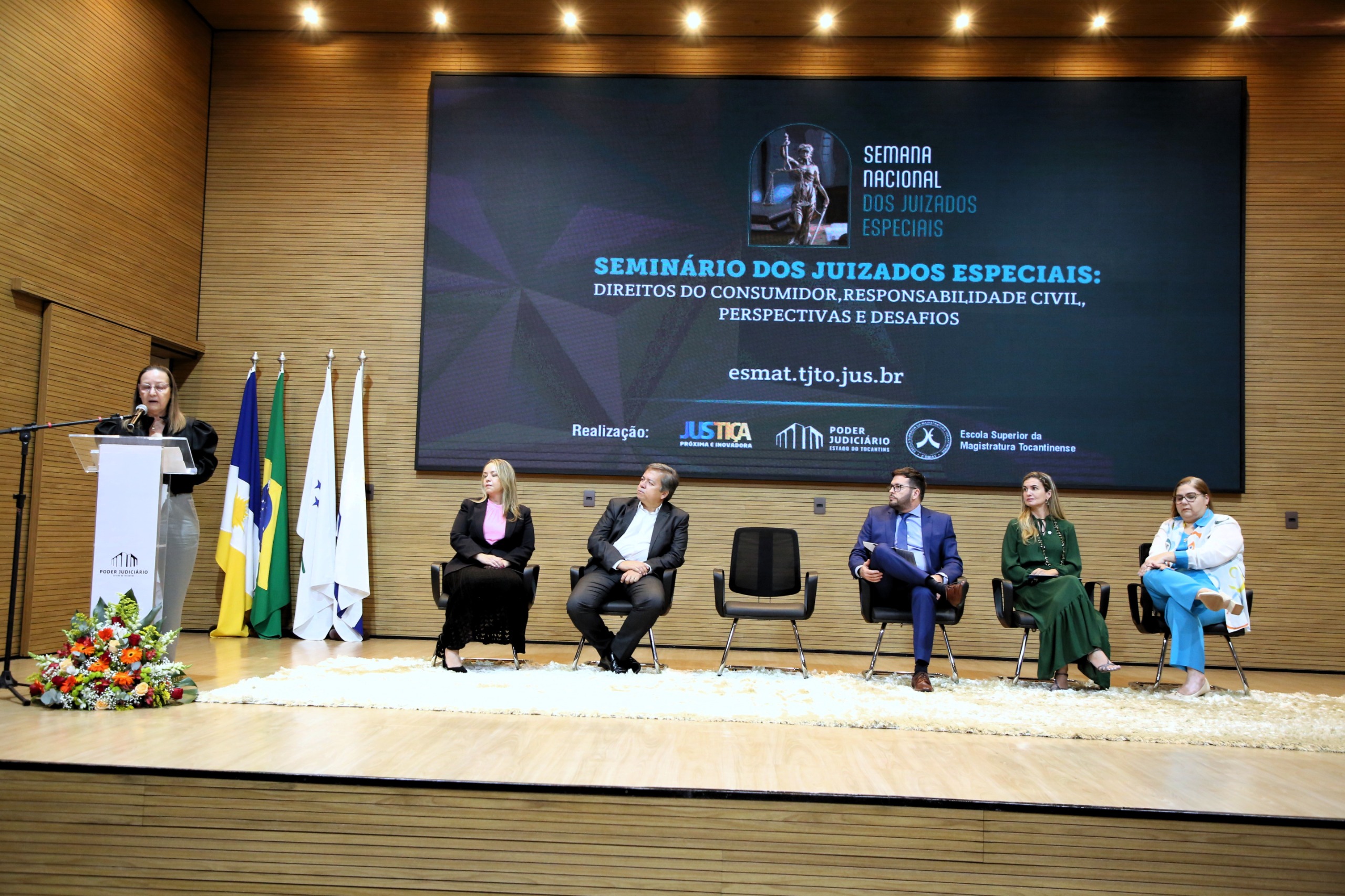 Desembargadora Maysa Vendramini fala ao microfone no púlpito à esquerda observada por autoridades que compõem o dispositivo de honra, três mulheres e dois homens, no palco do auditório do TJTO, durante a abertura do Seminario dos Juizados Especiais