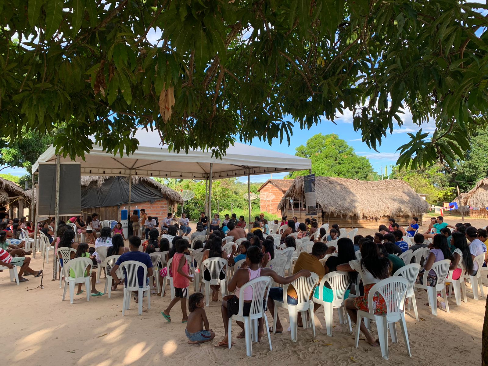 Imagem mostra pessoas sentadas em cadeira branca em baixo de uma tenda e de árvores