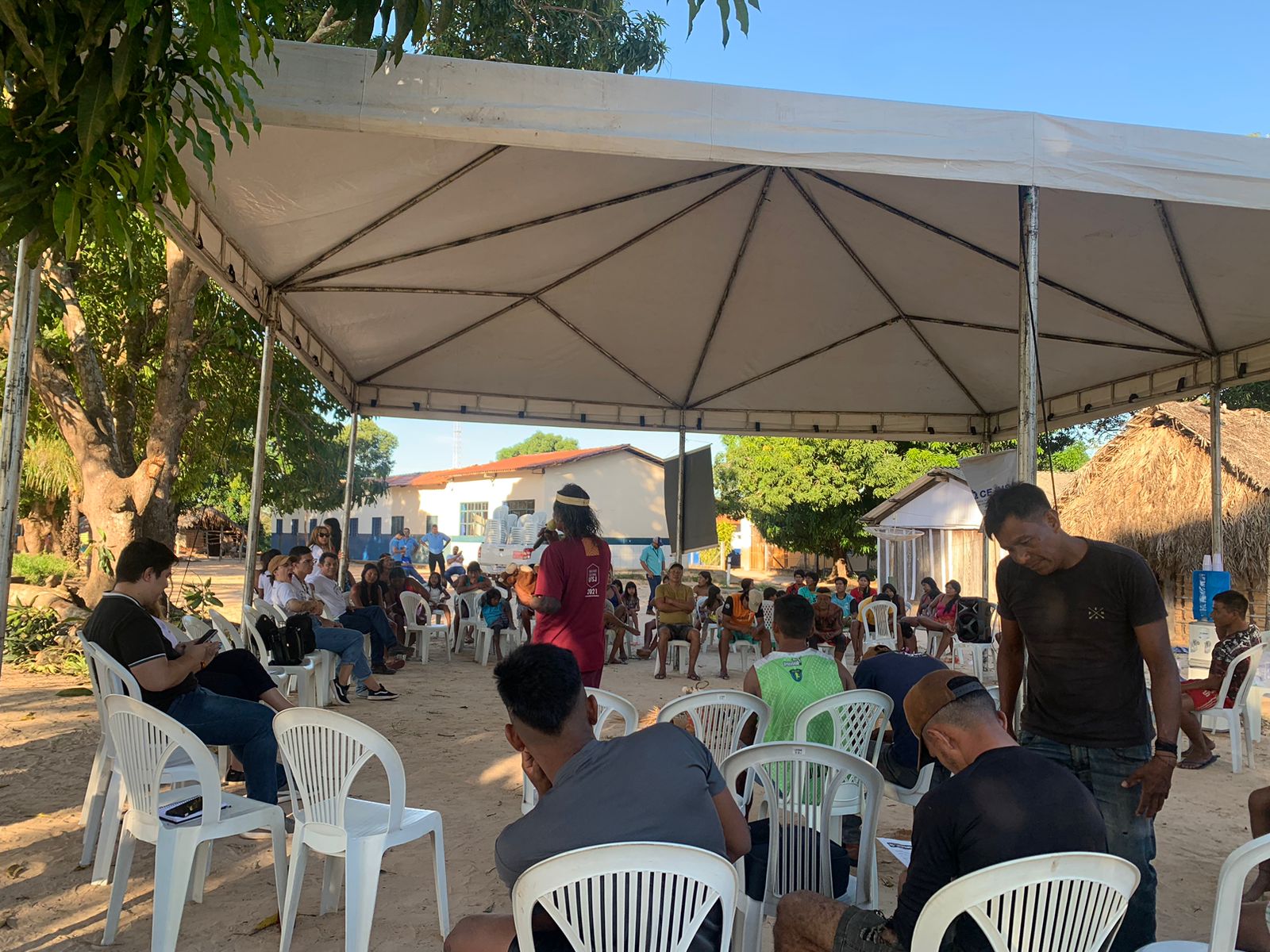 Imagem mostra pessoas sentadas em cadeira branca em baixo de uma tenda; ao centro uma pessoa fala com microfone