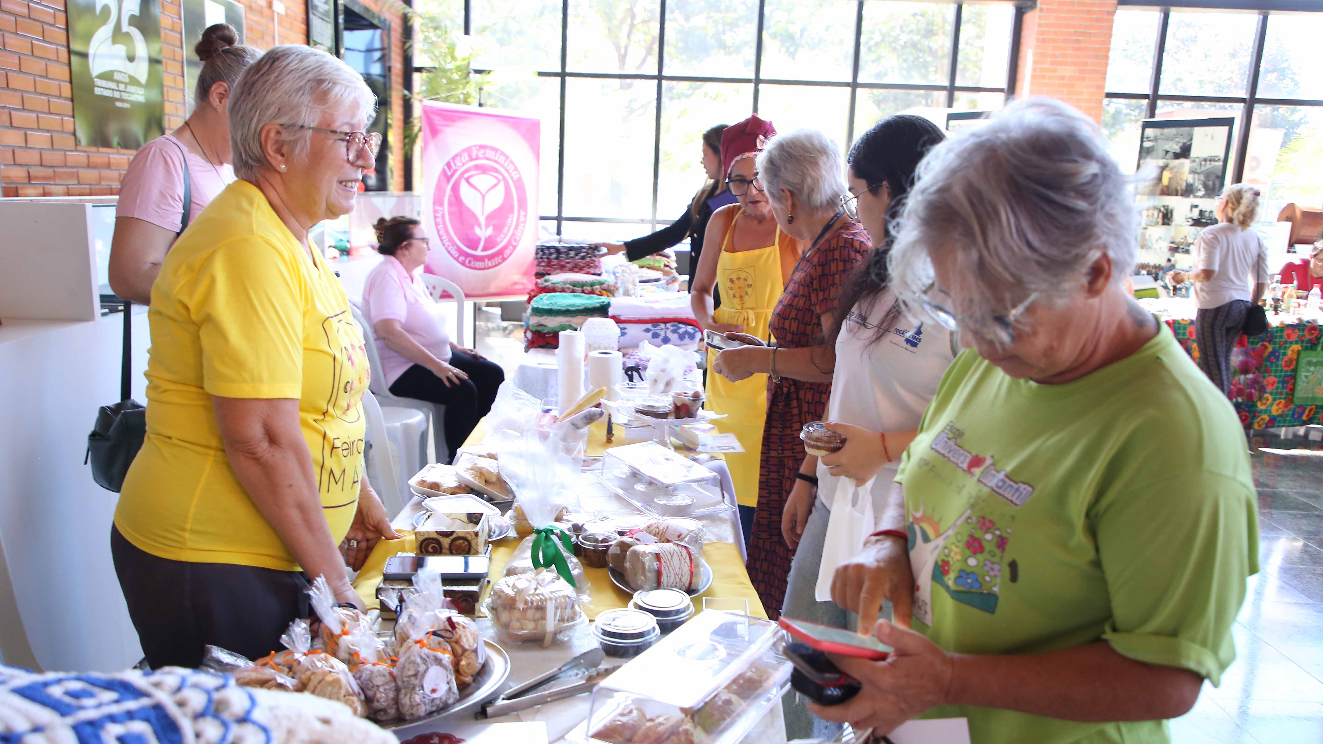 Fotografia colorida que mostra mulheres em pé expondo produtos de Feira 