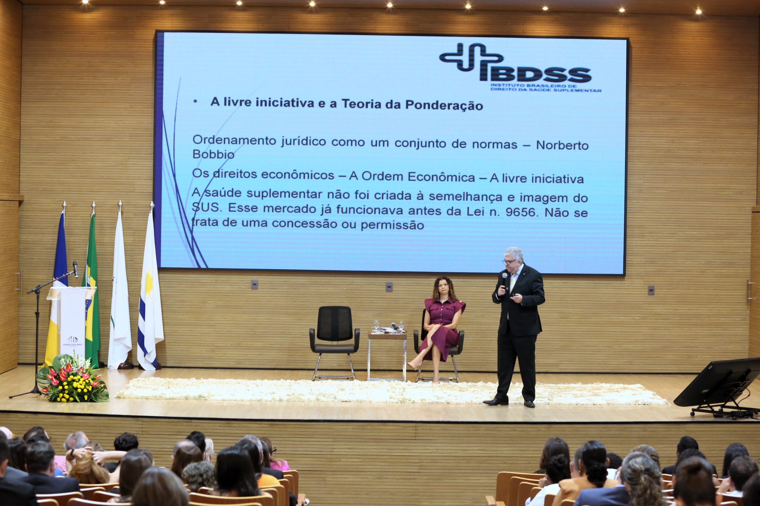 Imagem mostra um telão atrás e à frente pessoas em um palco ; uma em pé falando e a outra sentada