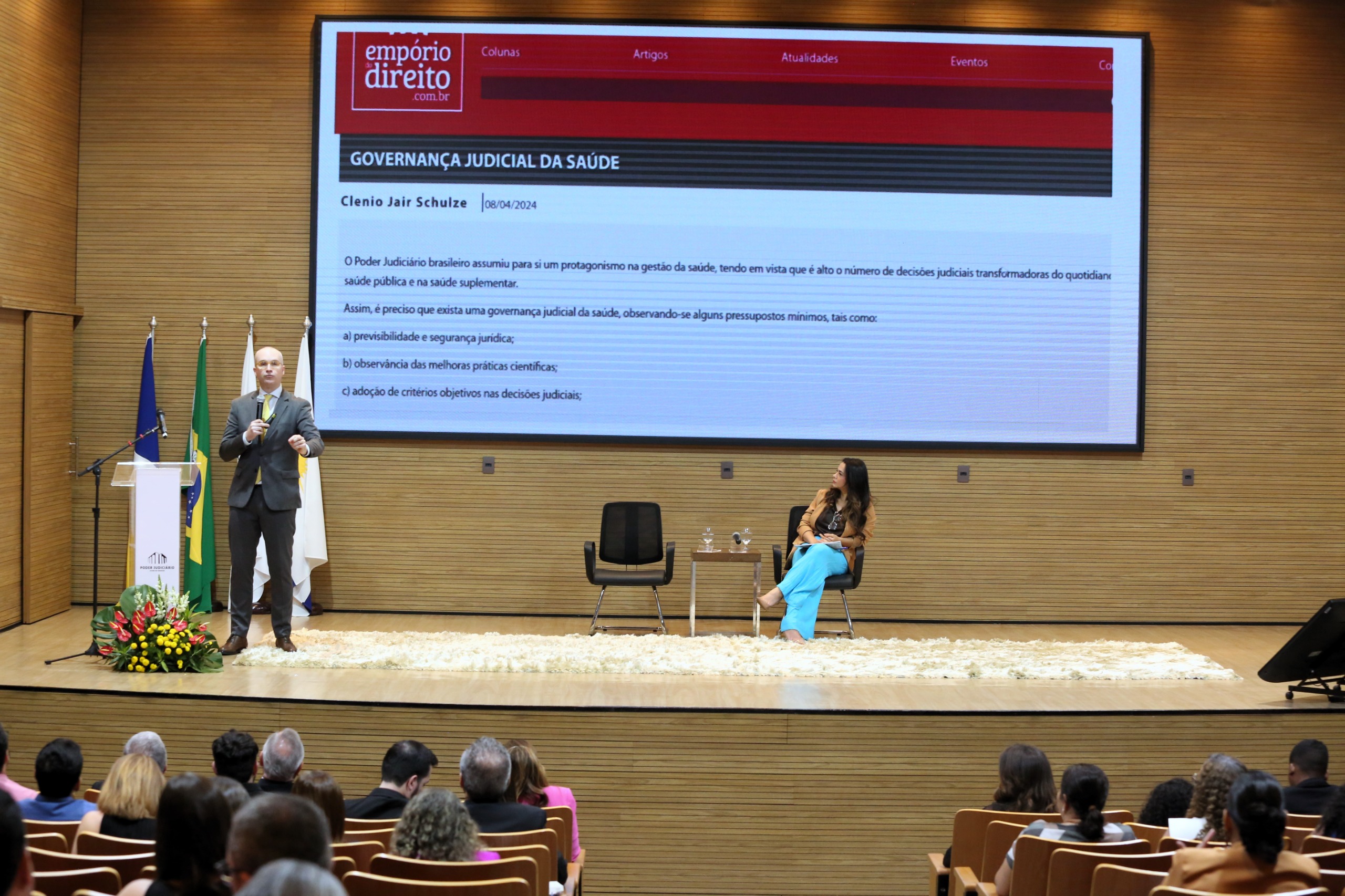 Imagem mostra um telão atrás e à frente pessoas em um palco ; uma em pé falando e a outra sentada
