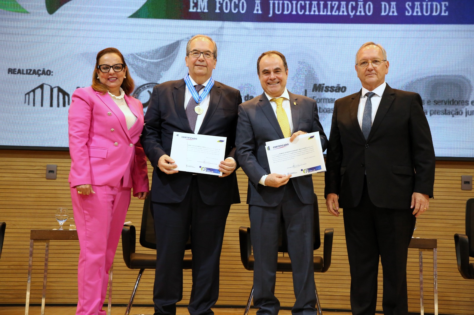 Imagem colorida que mostra três homens e uma mulher vestindo rosa, em pé, segurando um certificado 